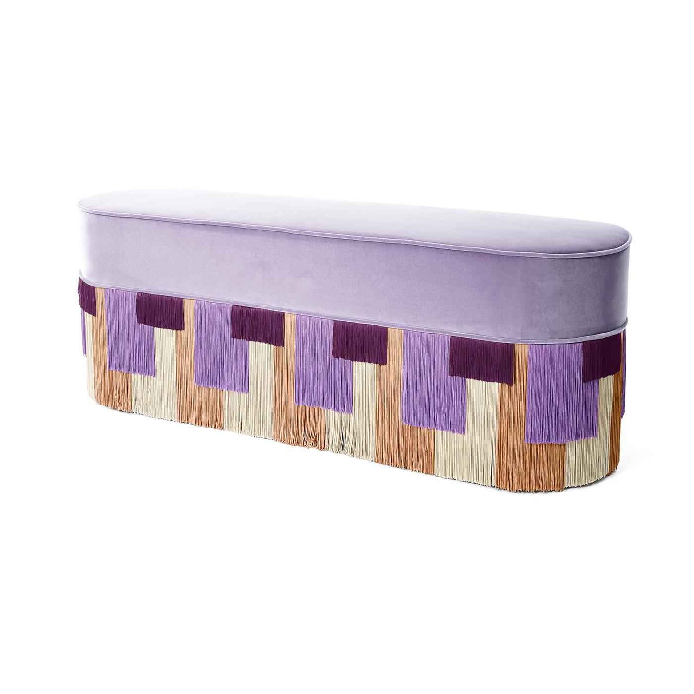 A long and classy pouf in a lilac color that has an array of fringes of different lengths and colors which appears as a sequence of colorful geometric shapes. The base is in painted beech wood and the top is comfortably padded and upholstered with