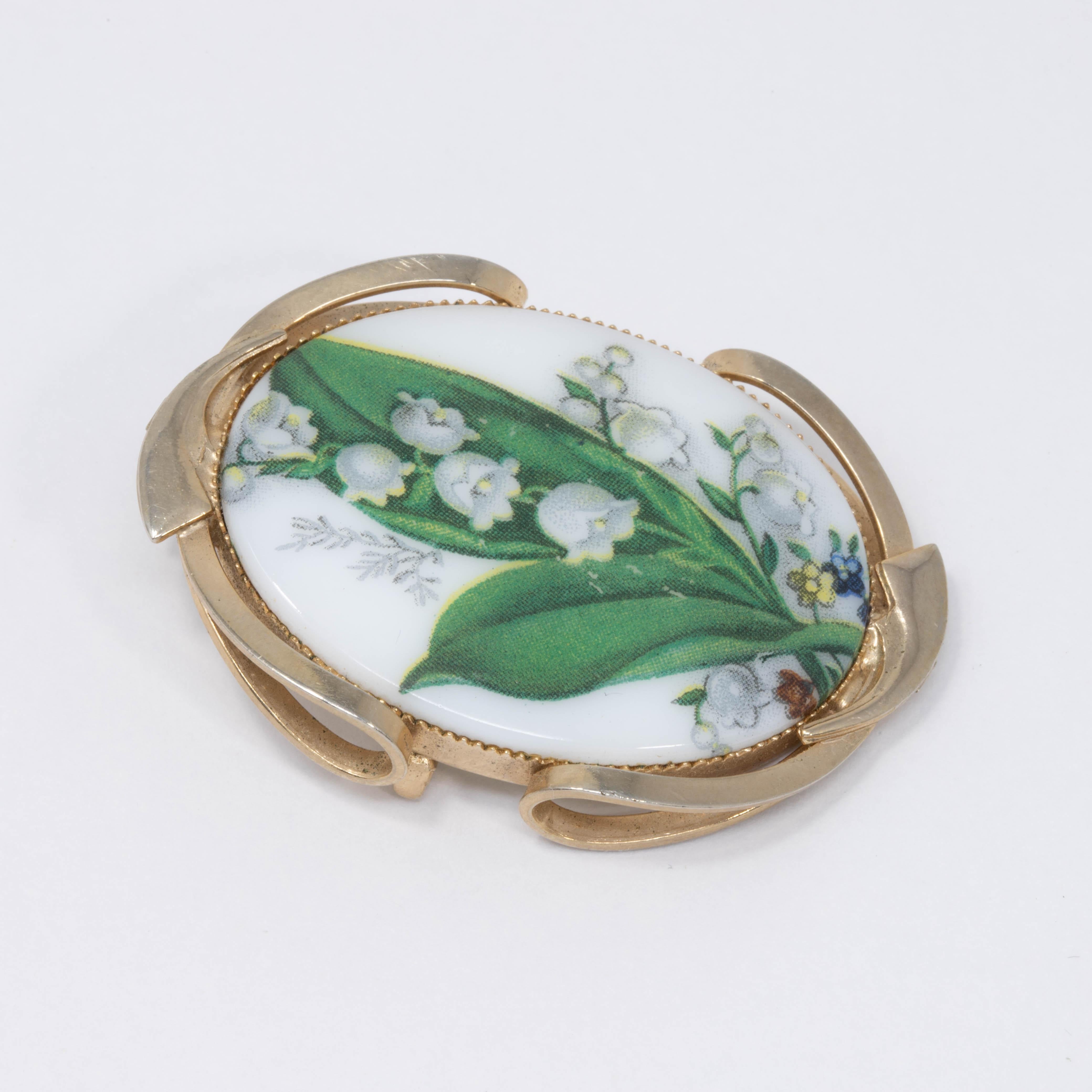 A lovely pin brooch and pendant! Features an oval white stone centerpiece with a painted lily of the valley, set in a decorative goldtone metal setting.
