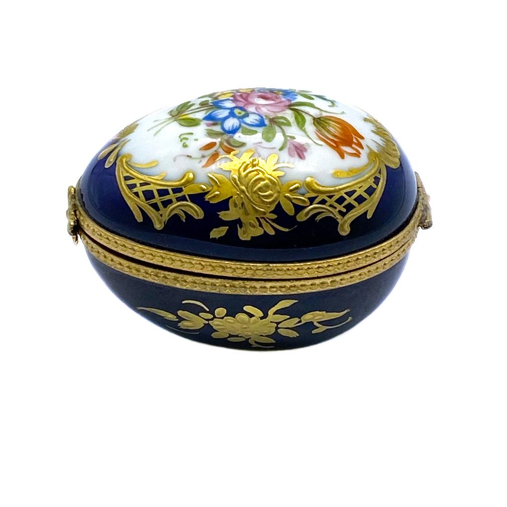 Il s'agit d'une boîte à bibelots en forme d'œuf de Limoges. Cette boîte en porcelaine bleu nuit est ornée de fleurs de style français peintes à la main et de décorations dorées. Il est doté d'une monture en laiton et d'un fermoir en forme de fleur.
