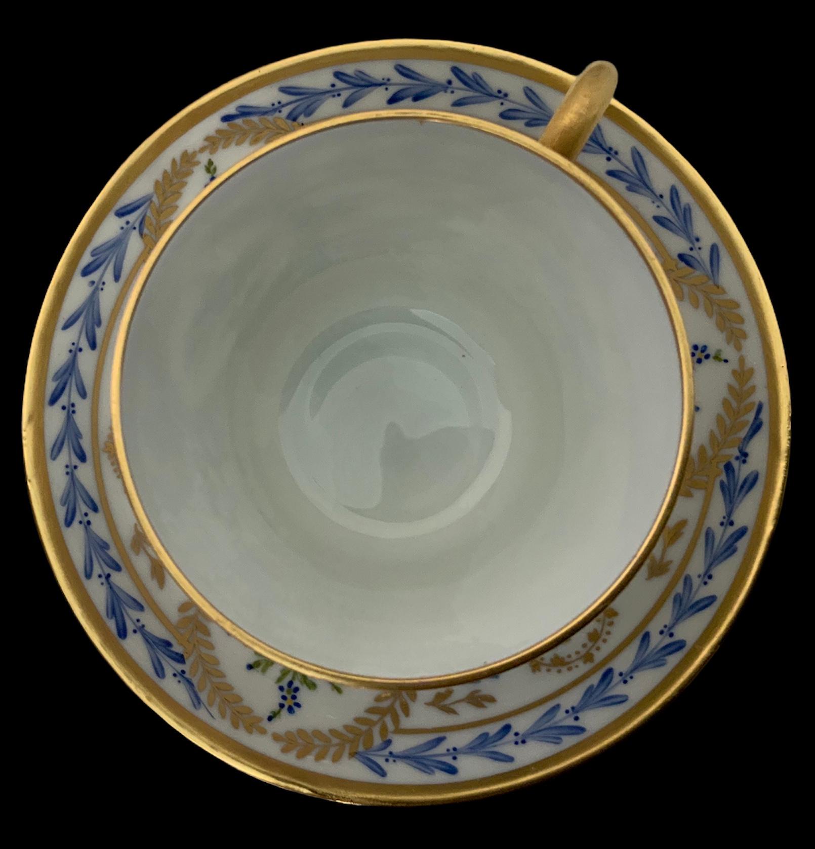 This a Limoges hand painted porcelain demitasse and saucer. It is adorned with a blue laurel garland alternated with a garland of gilt & blue leaves. In the center of it, there is some branches of blue forget me nots flowers surrounded by a gilt