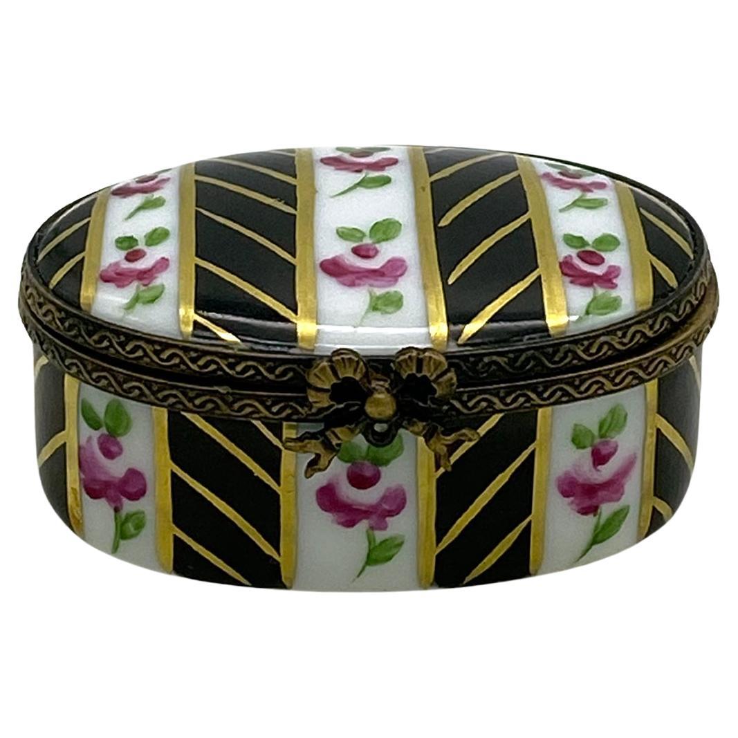 This is a hand-painted Limoges trinket box. This black background oval shaped porcelain box has pink flowers alternating with stripes. The brass hinge frame of the box has a ribbon clasp. This box is artist signed with 