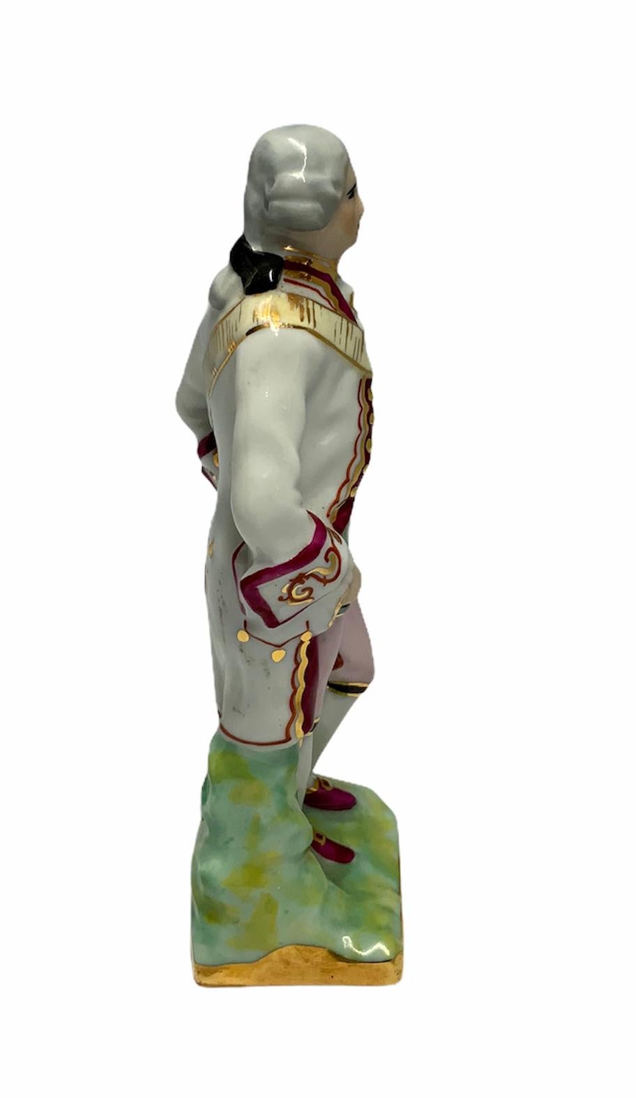 This a small Limoges porcelain depicting a man with an 18th century gentleman suit. He is standing in front of some green bushes. Under the base is the Limoges porcelain hallmark.
