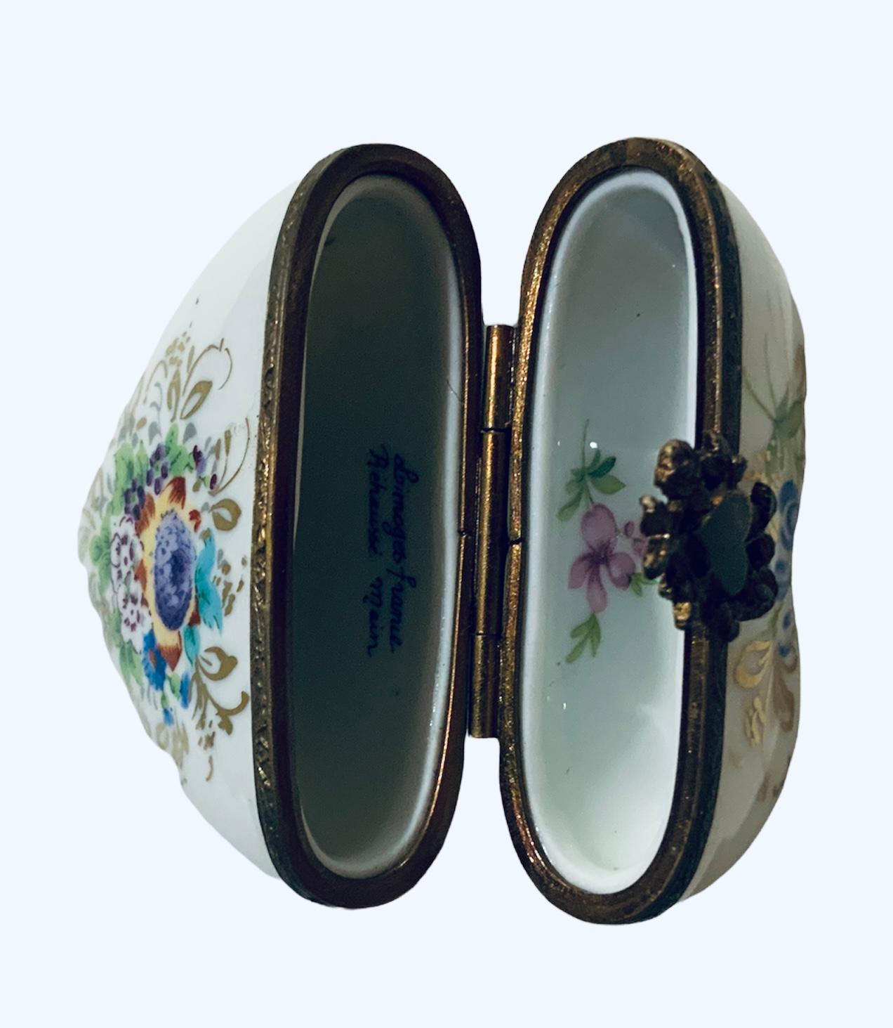 This is a Limoges France porcelain hand painted hinged trinket box. It is adorned with different bouquets of flowers with gilt branches around it and inside the lid. The clasp is a bronzed brass heart surrounded by foliage. It is signed Limoges,