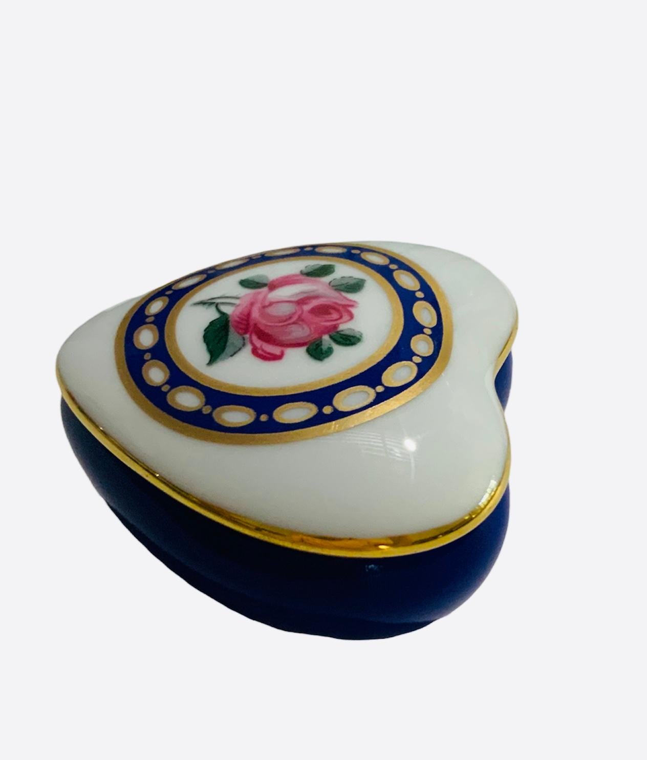 This is a small Limoges France porcelain hand painted lidded trinket box. It is adorned with a rose in the center of its white lid and a gilt royal blue wide circlet with gold rings. Also, there is another rose inside the box. The bottom of the box