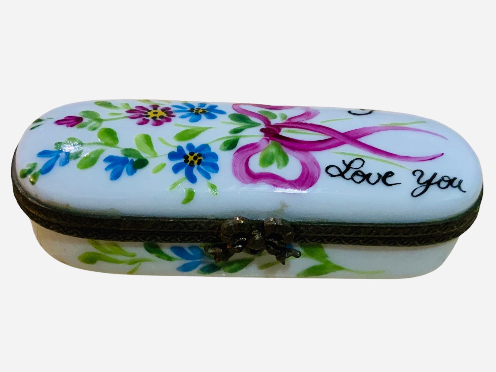 Se trata de una caja de baratijas de agujas de porcelana de Limoges Francia. Su fondo es blanco. Está pintada a mano y adornada con un ramo de flores sujeto por un lazo largo rosa. También junto al ramo está escrito 
