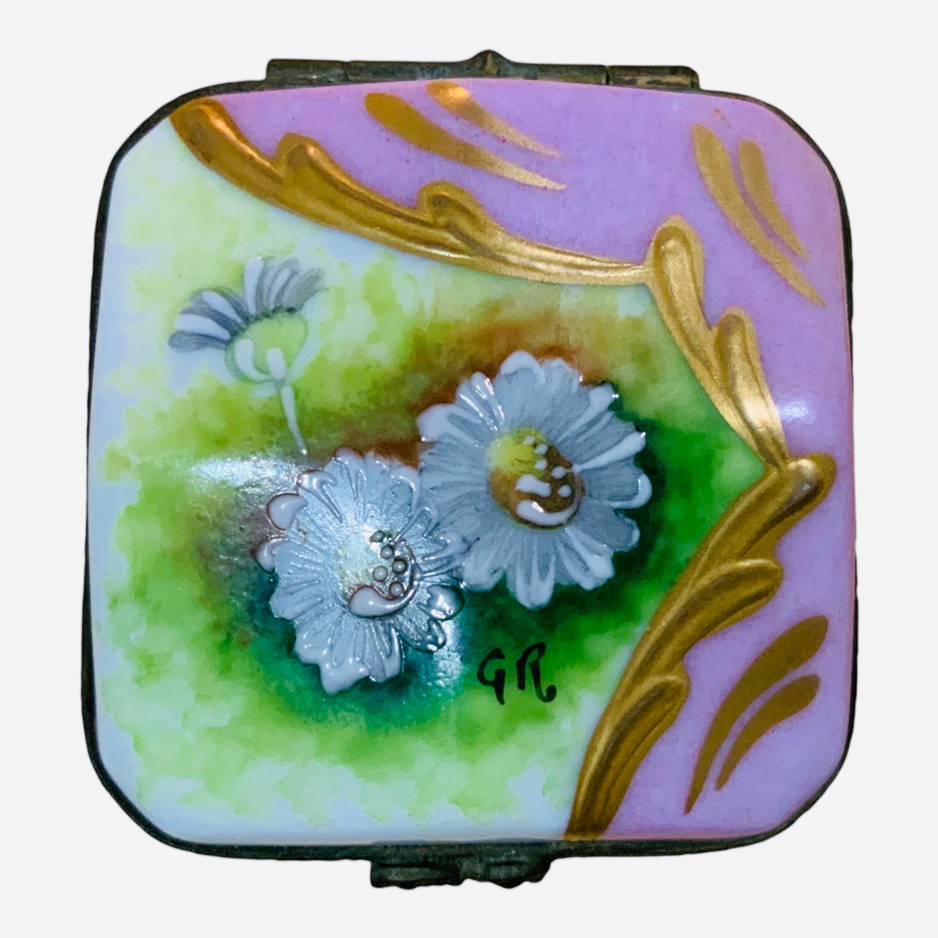 This is a Limoges Porcelain Octagonal Trinket Box. Its background is hand painted pink and white. The top of the hinged lid is adorned with a green grass with three daisies. Some gilt paint brushes framed it as well as the base of the box. The front