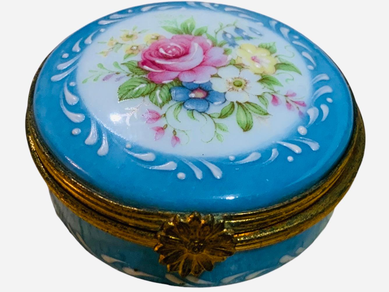 Se trata de una caja redonda de baratijas de porcelana de Limoges. Su fondo es azul cielo pintado a mano. La parte superior de la tapa abatible está adornada con un ramo de flores de colores con ramas verdes alrededor. El interior de la tapa está