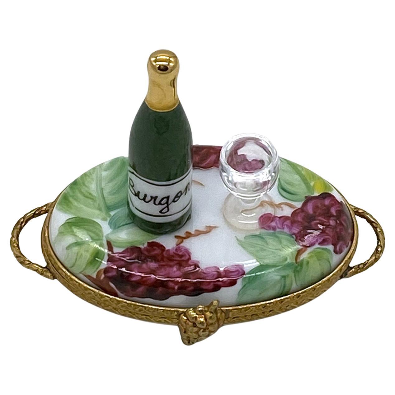 This is a hand painted Limoges trinket box with wine bottle and glass. A bottle of burgandy and a wine glass set on a oval shaped tray forms the lid ofthe box. The porcelain tray box is painted with ruby grapes and leaves, also with 