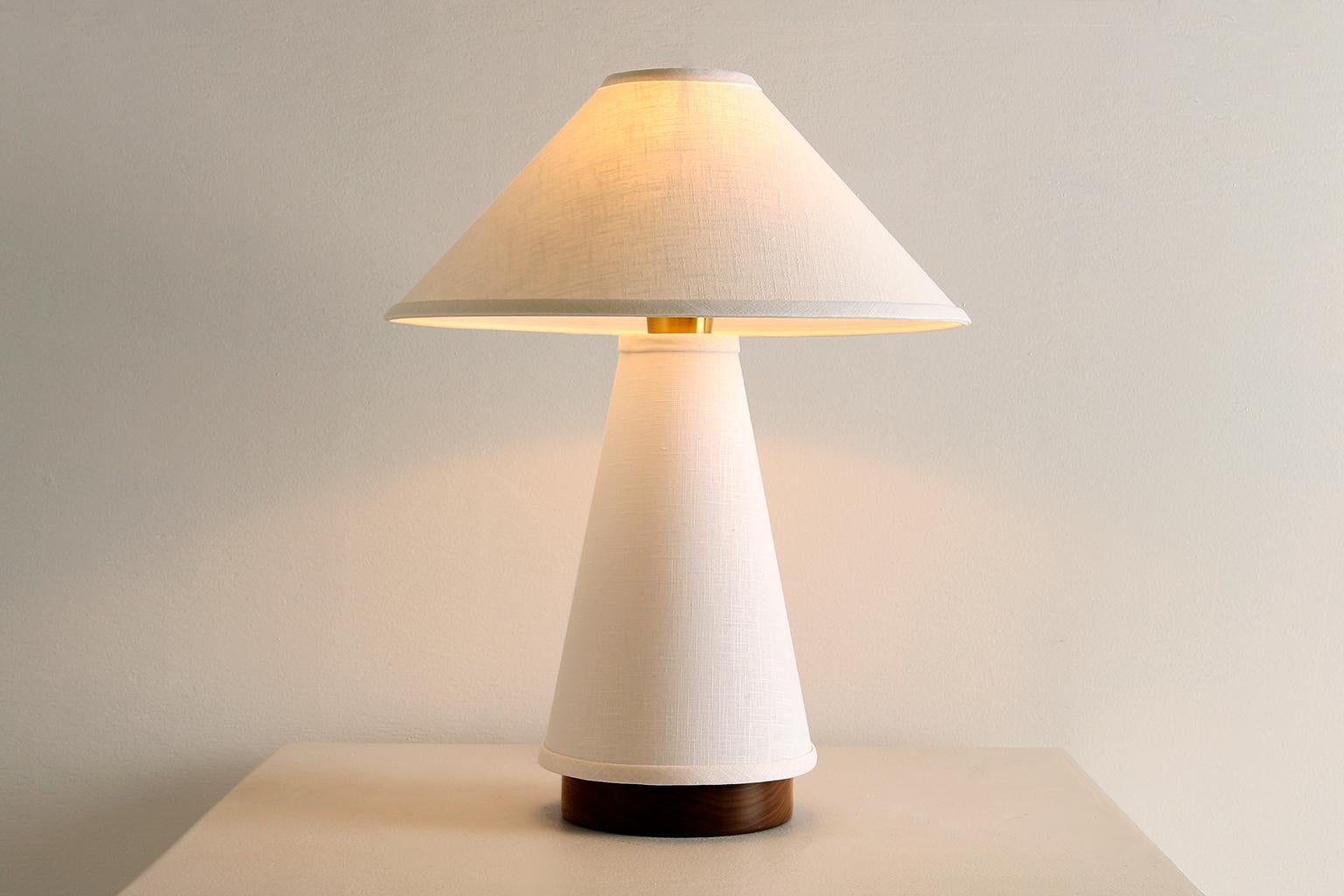 La lampe de table Linden se caractérise par un abat-jour et un corps de lampe en lin crème, une base en noyer massif et des détails en laiton. Étude de la dualité, la pièce explore l'équilibre entre les tissus souples et fluides et les formes