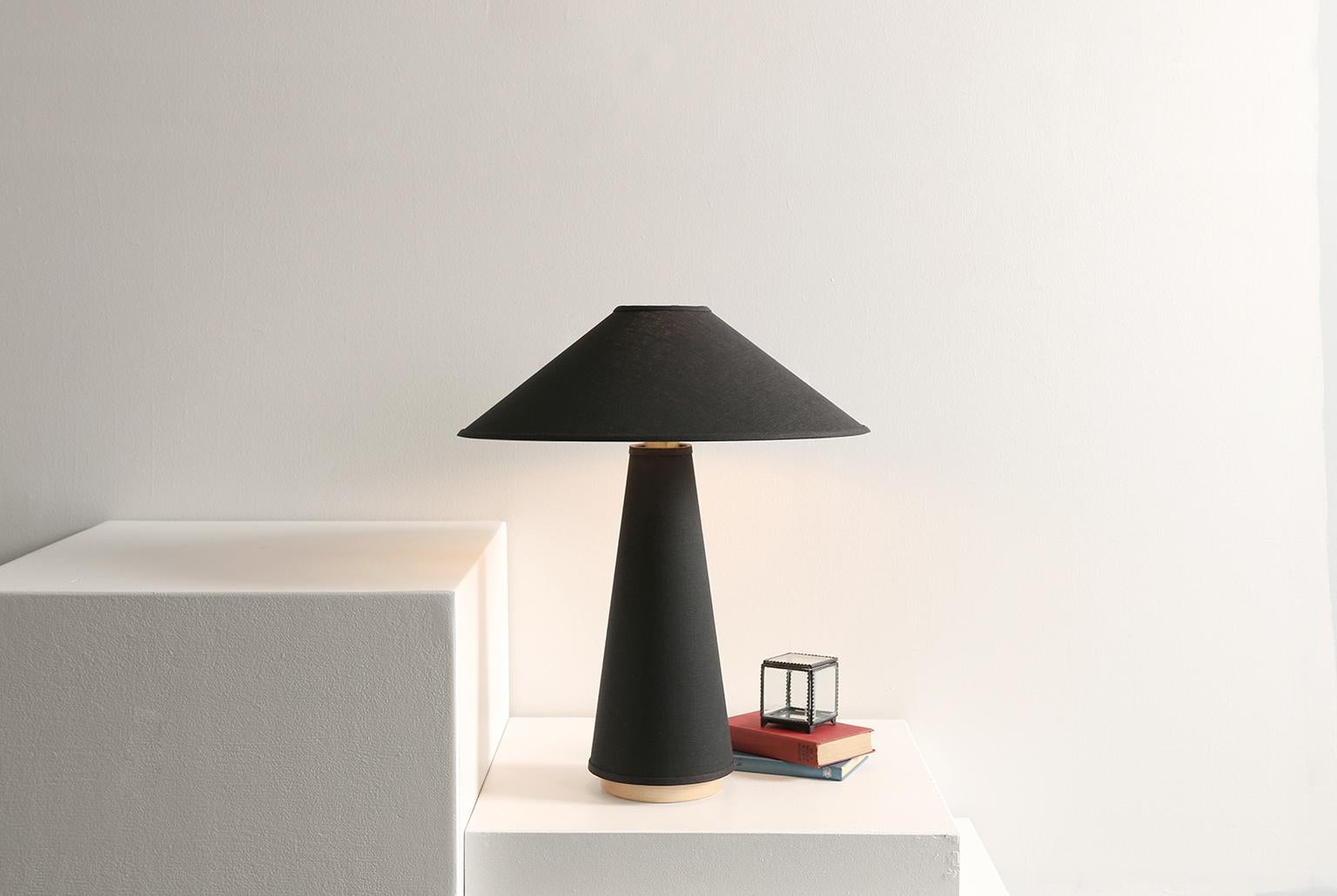 The Linden table lamp features a black linen shade and lamp body, solid hardwood maple base, and brass details. The tabletop fixture references the Classic profiles and angles of Mid-Century Modern design with distinct clean lines, smooth sweeping