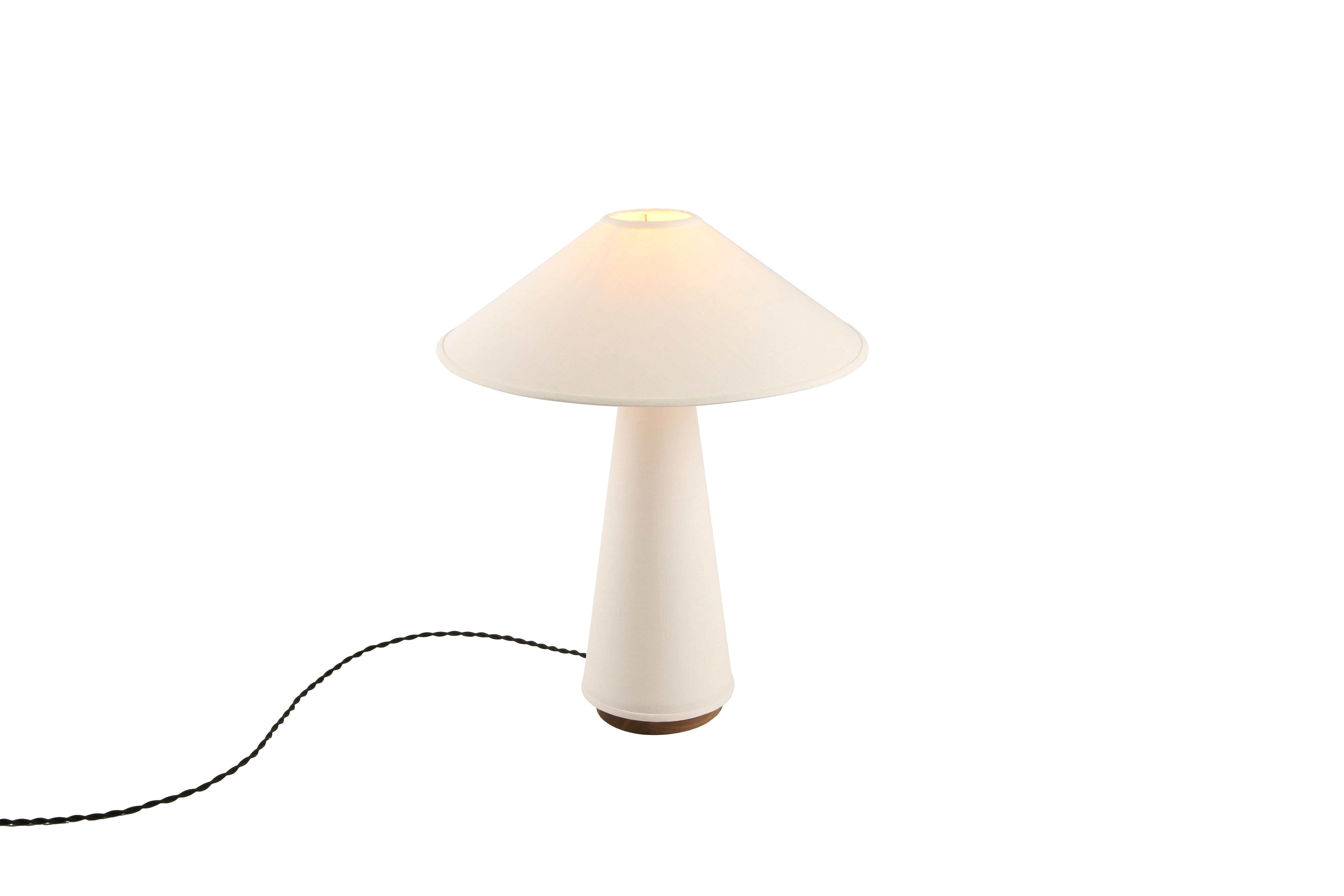 La lampe de table Linden présente un abat-jour et un corps de lampe en lin crème, une base en bois dur massif de noyer et des détails en laiton. Étude de la dualité, cette pièce explore l'équilibre entre un tissu souple et fluide et des formes