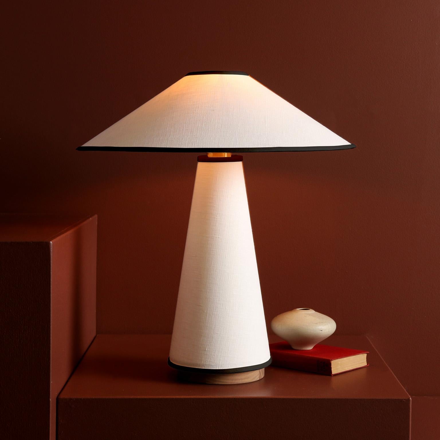A contemporary table lamp with a linen shade and body, hardwood base, and brass details. The structural shape, distinct clean lines, and neutral tones are derived from classic mid-century modern design. Linen fabric gives a lustrous, glowing quality