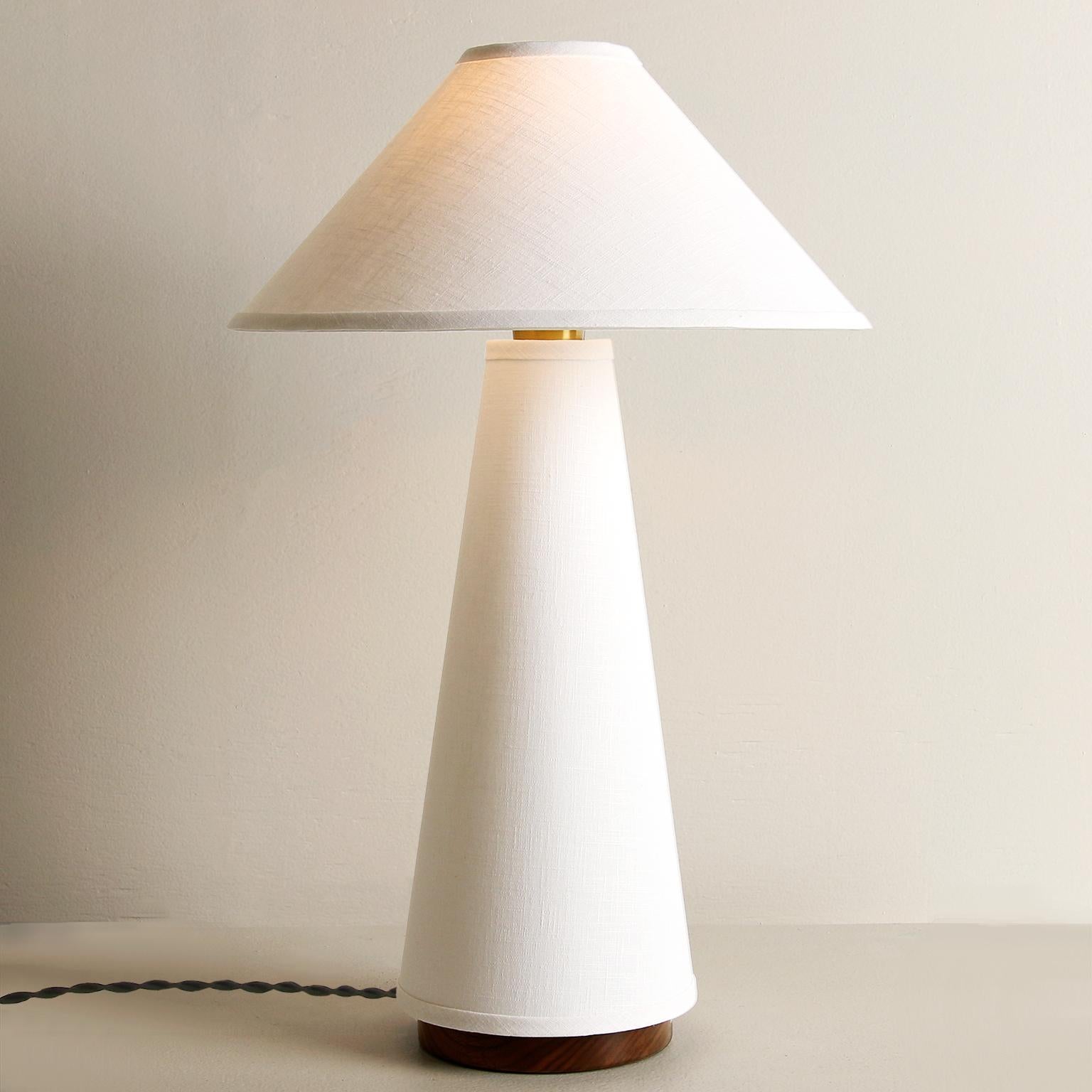 The Linden table lamp features a cream linen shade and lamp body, solid hardwood walnut base, and brass details. A study in duality, the piece explores the balance between soft, flowing fabric and structured, conical forms. Bulb sold separately.