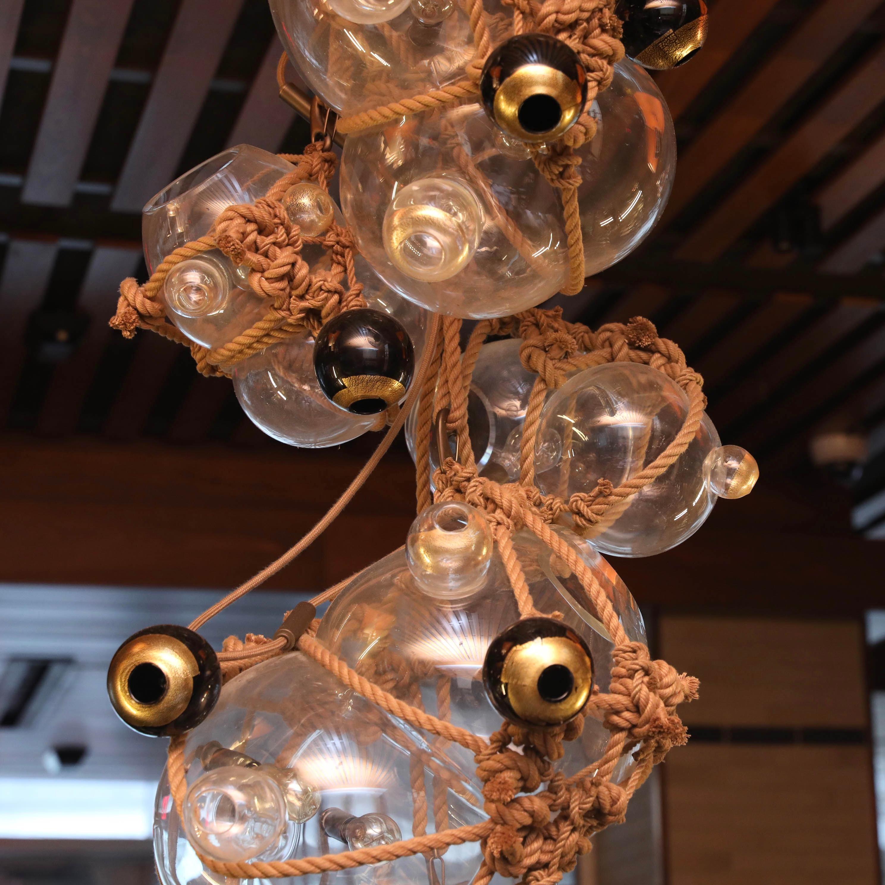 Inspired by Japanese packaging, buoys, and shibari, knotty bubbles is a sculptural light made of hand-blown glass 'bubbles' bound together with knotted rope. This chandelier is a combination of two stacked knotty bubble chandeliers. Each of the