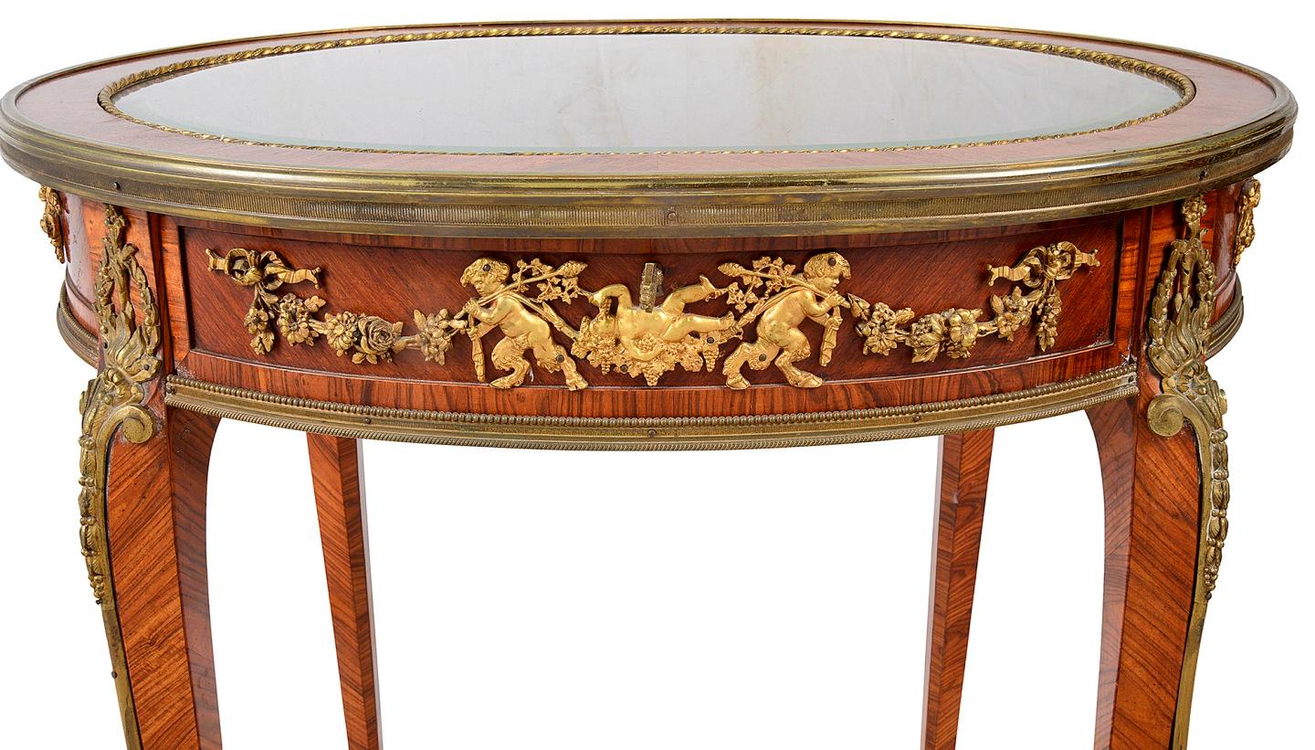 A fine quality late 19th century mahogany oval table display cabinet, having wonderful gilded ormolu mounts depicting cloven hoofed Bacchus influenced putti playing amongst grapes, vine leaves and garlands of flowers. The hinged glazed lid opening