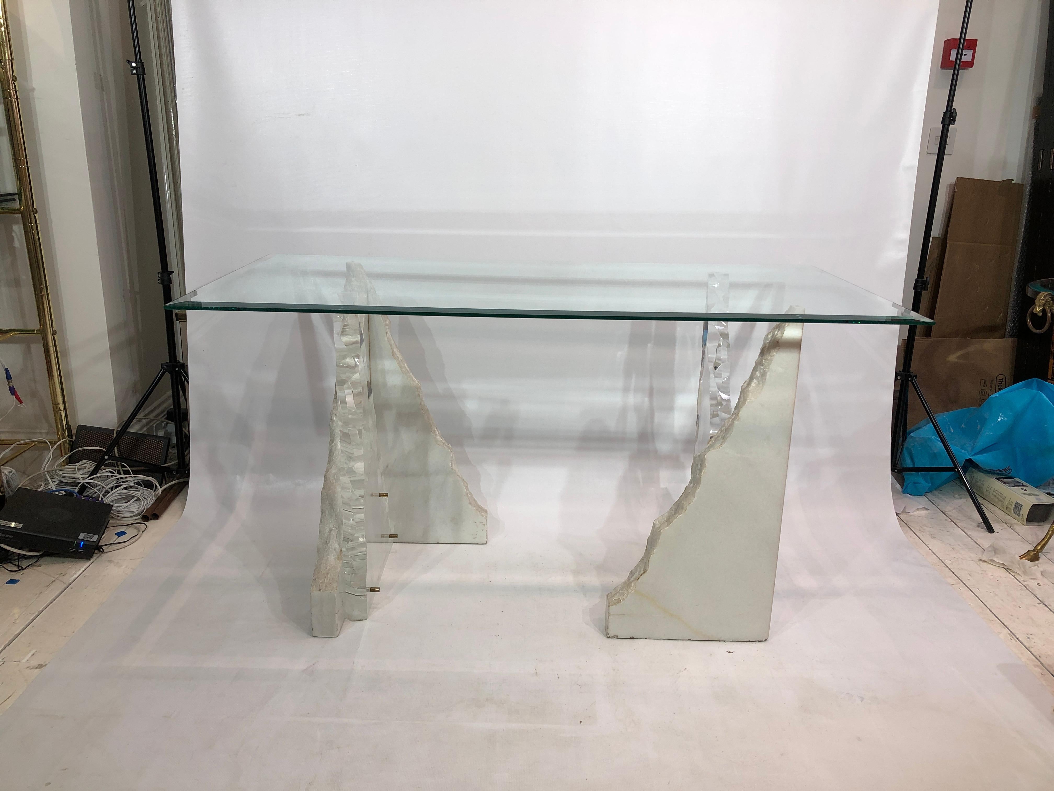 This striking glass top desk or table, manufactured by popular 1970s acrylics company Lion in Frost, features two thick lucite and raw edge carrara marble corner pieces with brass fixings, strongly resembling an iceberg or stalagmite. A sheet of