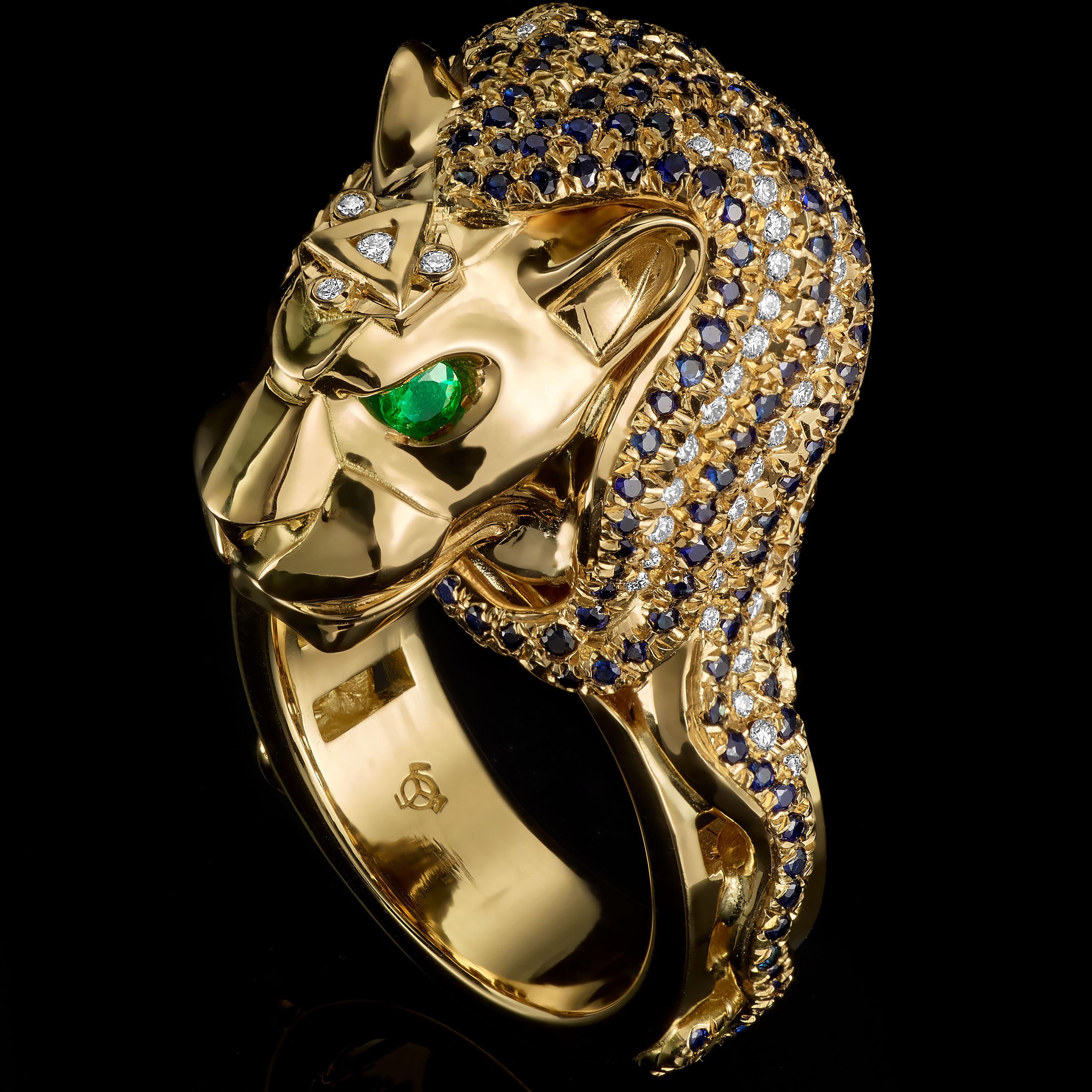 Lion ring in 18K gold set with diamond and sapphire pave and emerald eyes. A unique, high end jewel destined to make an impression. 
A bust of a lion sits proudly atop a geometric ring with faceted sides. Its mane of blue sapphires and diamonds