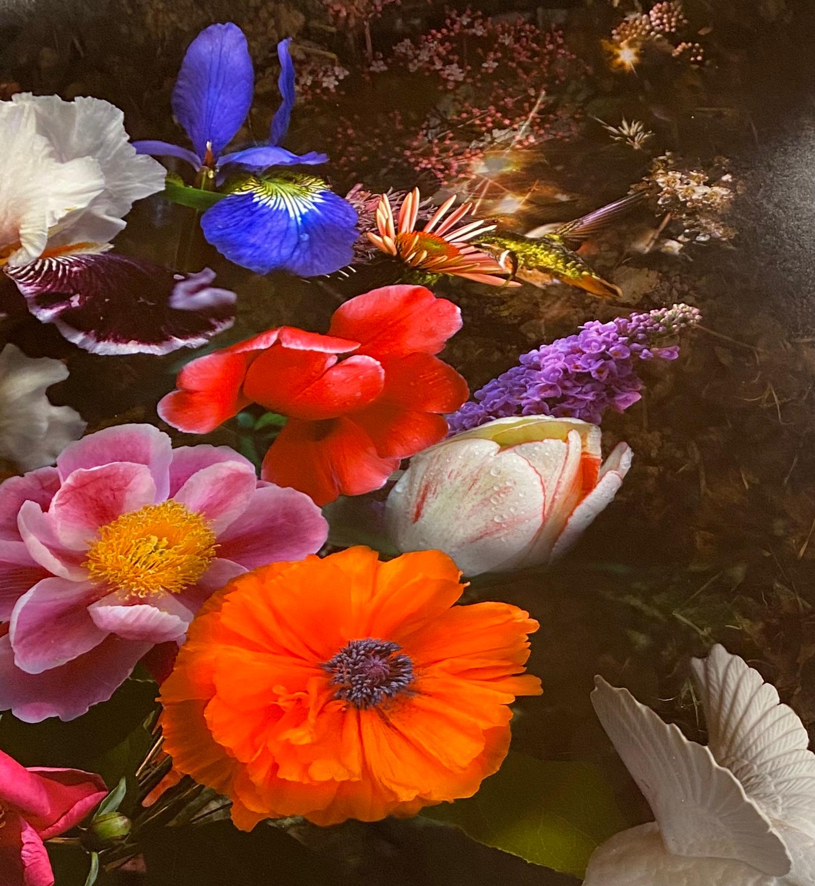 This botanical still life arrangement of orange, pink, yellow and purple flowers is dramatic and mysterious against a dark black background. A white porcelain bird figurine stands to the right of the bouquet, a crystal geode and shell to the