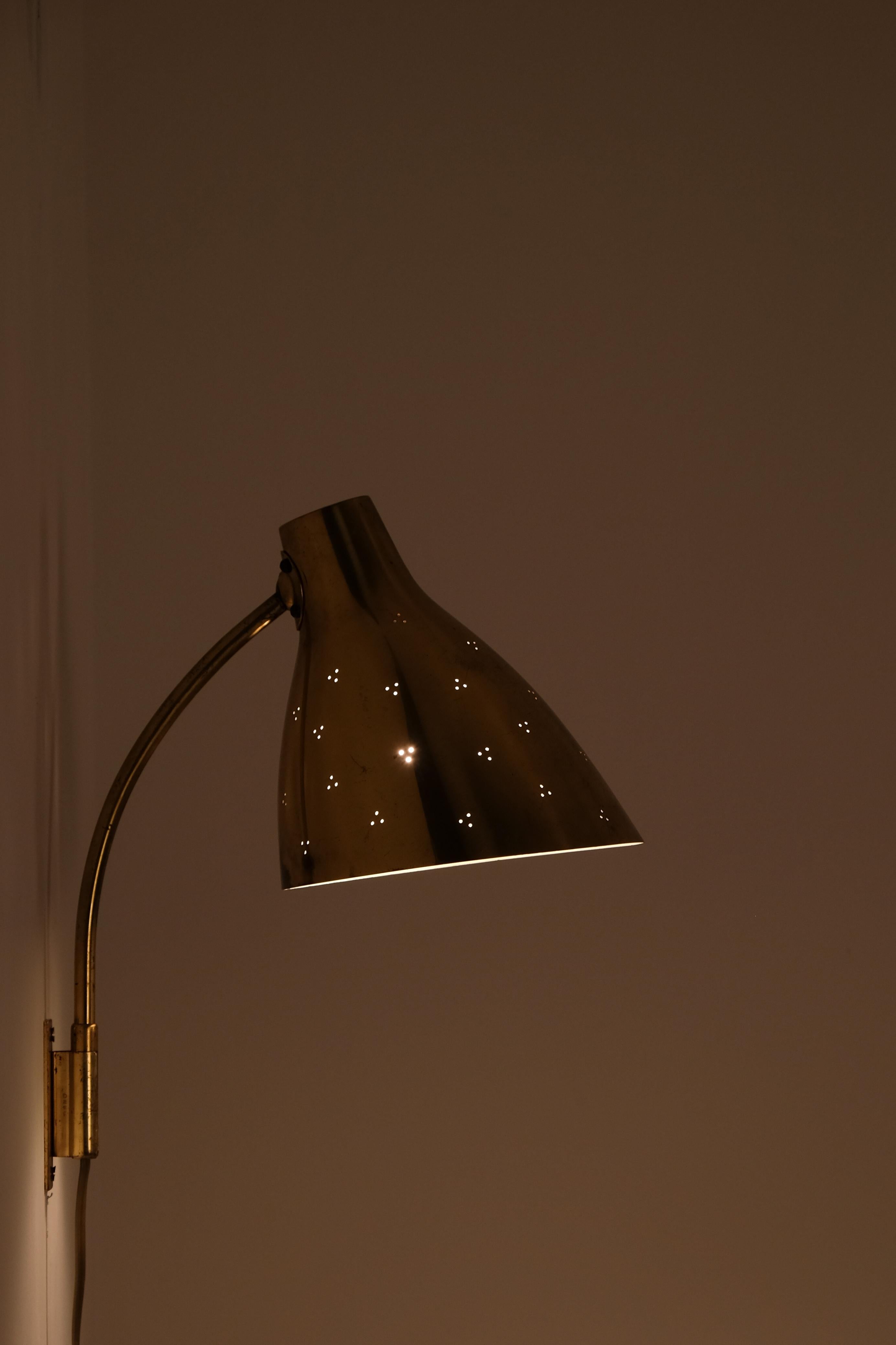 This perforated brass wall lamp model 50-055 designed by the renowned Finnish designer Lisa Johansson-Pape. With its elegant curved arm and beautifully perforated shade, this lamp emits a soft, atmospheric glow while adding a touch of timeless