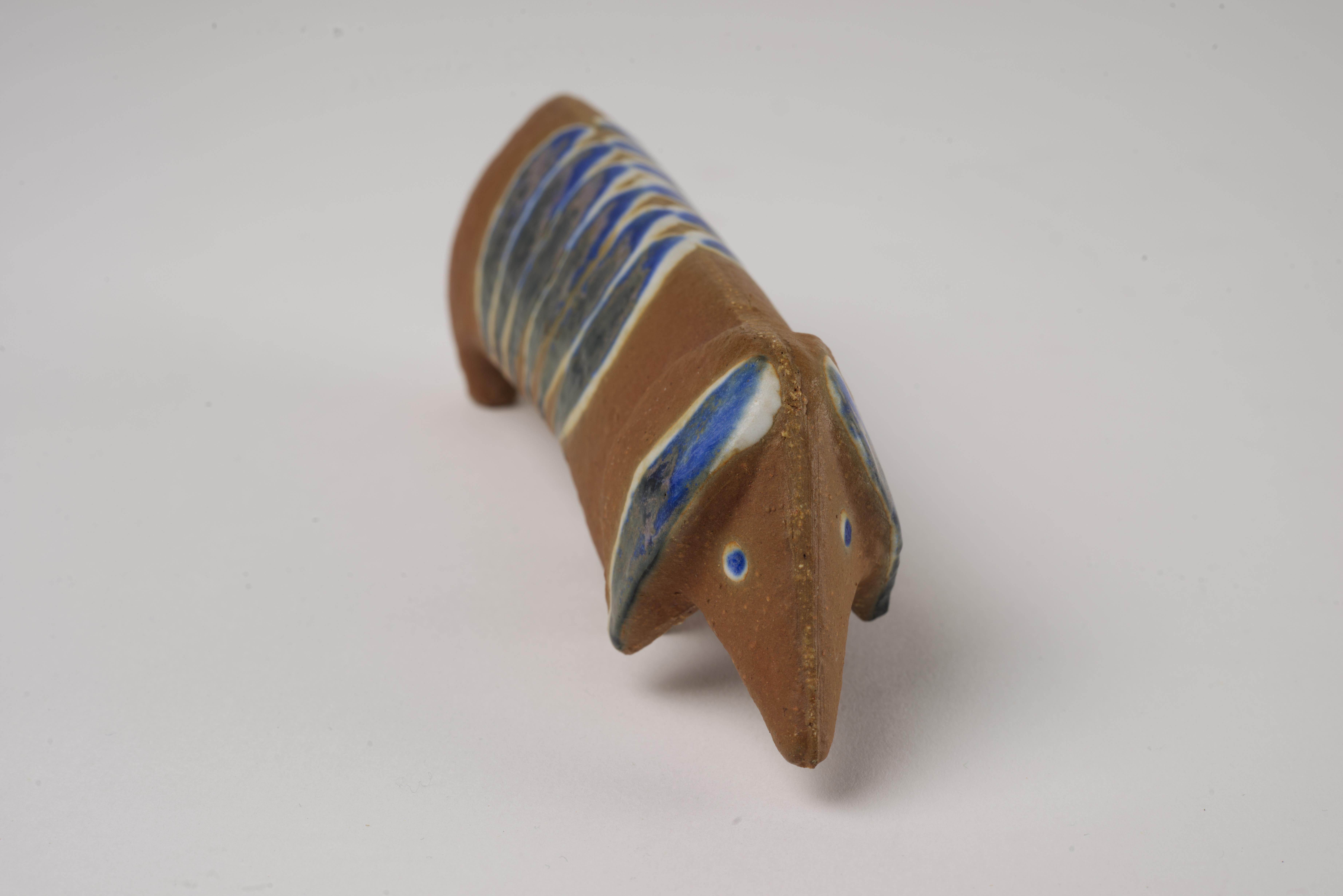  Dachshund dog is made of chamotte stoneware with geometric decoration in blue and white semi-matte glaze. It was designed by Lisa Larson (1931-2024) in 1955 as part of the ”Lilla zoo” (Small Zoo) series for the Gustavsberg Porcelain Factory. 

The