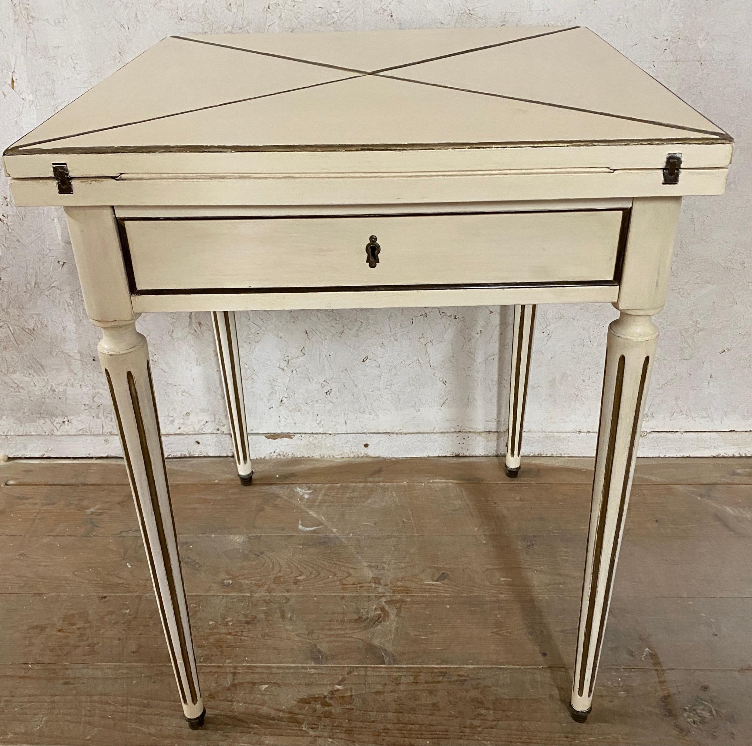An elegant French envelope game table. By opening the four hinged triangular sections the small end table in the neoclassical style becomes a square game or card table with black leather, gold embossed playing surface. Painted white, the table has