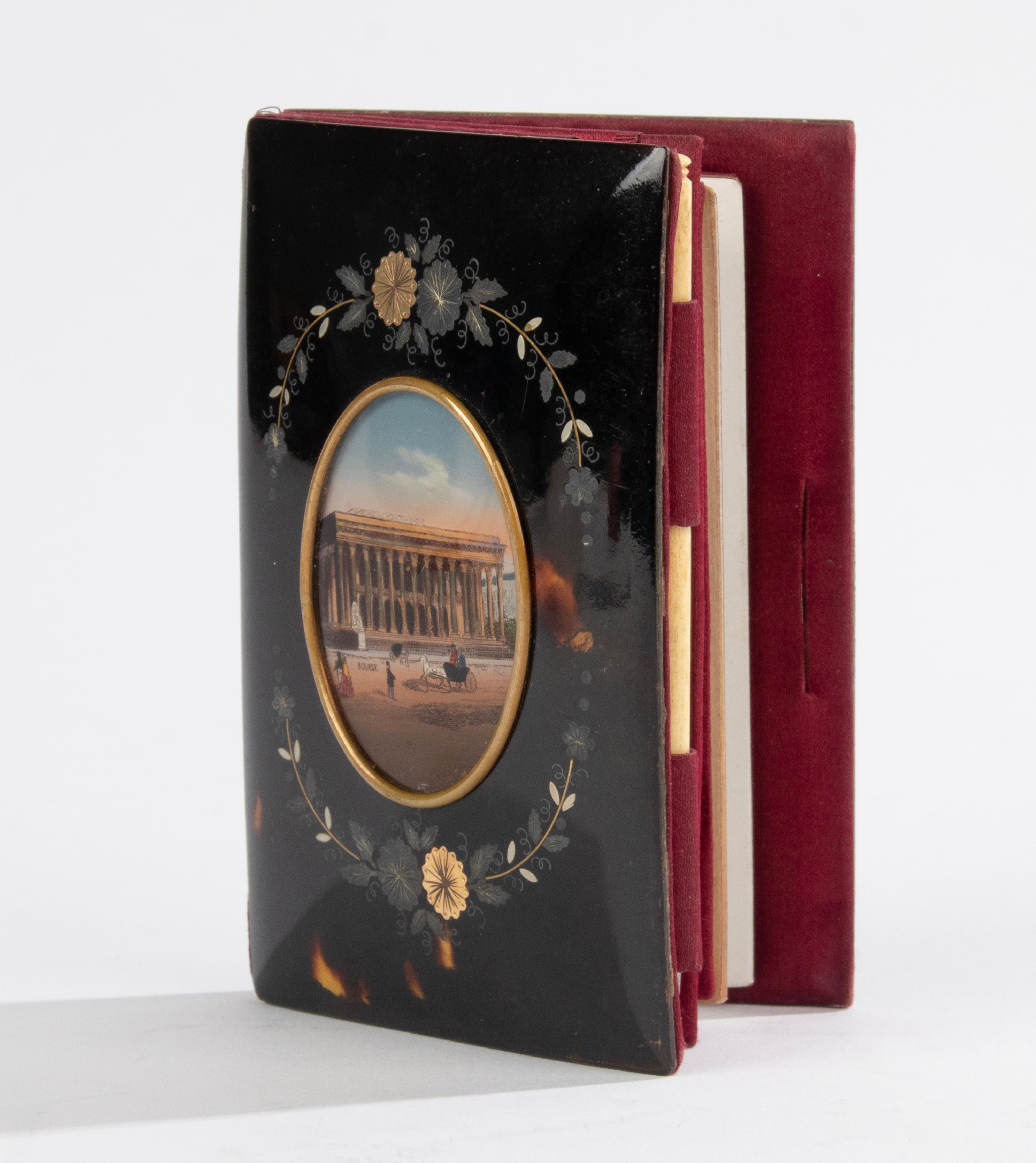 A small antique notebook. At the front cover is a hand painted medallion depicting the Palais Bourse building, the Paris stock exchange. Surrounded with foliage of tin and copper inlay. The cover is made of faux tortoise, a synthetic