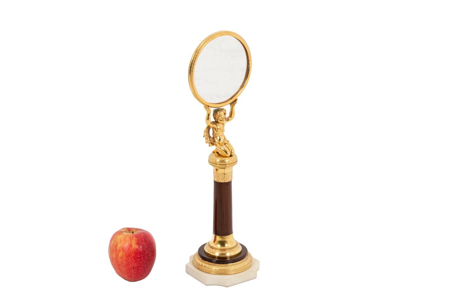 Petit miroir de forme ovale en bois de rose et bronze doré reposant sur une structure tubulaire. Cadre en bronze doré. Figure d'un enfant à genoux soutenant le miroir avec ses deux bras épargnés. Base moulée en bois et bronze ciselé et doré. Socle