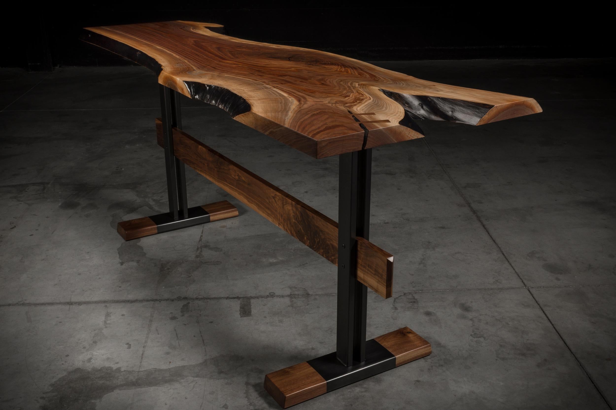 Der Cadieux-Tisch besteht aus einer einzigartigen massiven Nussbaumplatte mit lebendiger Kante, die auf einem eleganten Stahlsockel ruht. Der Sockel ist außerdem mit massivem Nussbaumholz verstärkt, um sowohl strukturelle Unterstützung als auch Stil