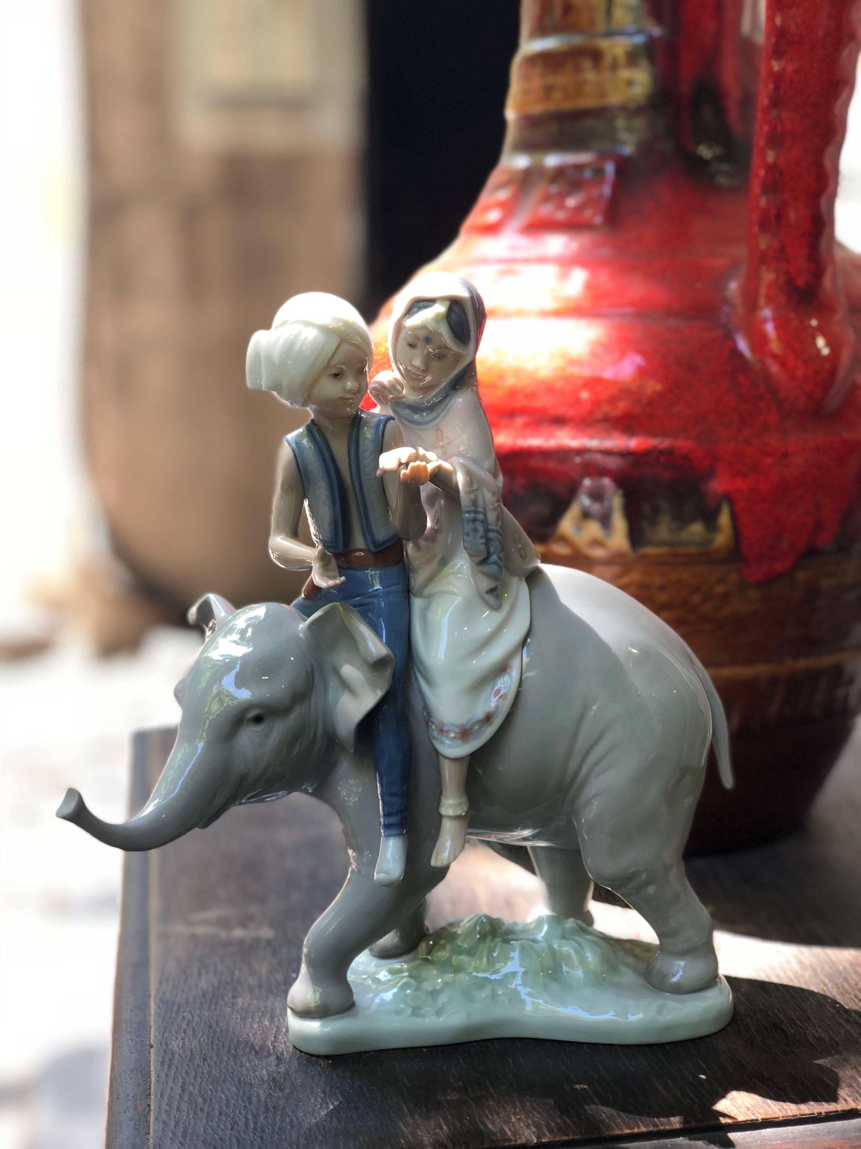 This Lladro piece is a porcelain figurine that depicts an young Indian elephant, upon which a pair of Hindu children sit. The glazed grey hues of the elephant's porcelain skin accentuates the intricate detailing and musculature of the largest land