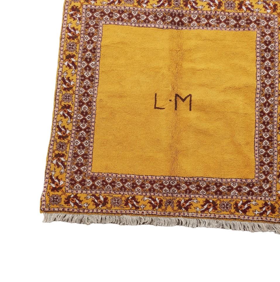 This is a square rug of great sophistication, with a minimalist yet elegant design. The main field of the carpet is a uniform and warm ochre/mustard color that exudes a cozy and comfortable feeling. In the center of the rug is the monogram 