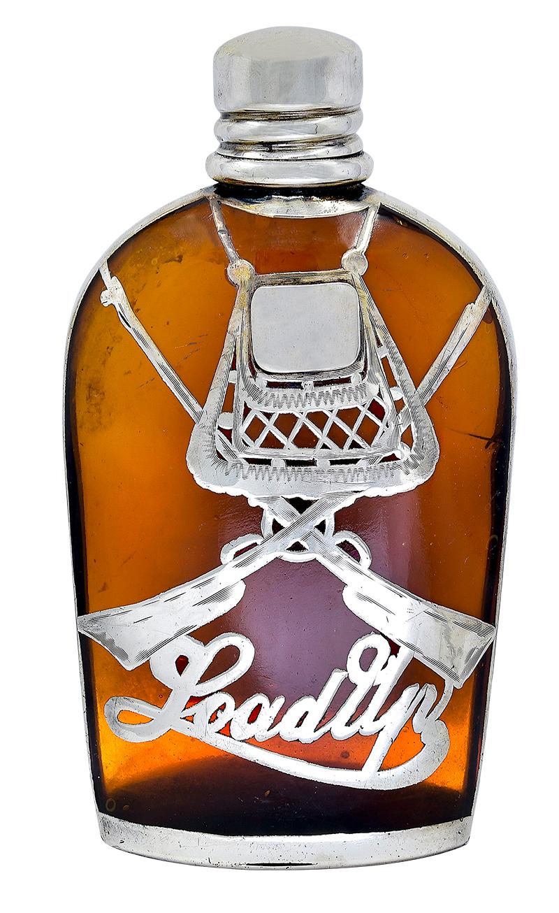 An antique American amber-colored glass flask with overlay of pierced sterling silver.  The front of flask features a pair of figural rifles in crossed form.  Under the guns, sterling silver script letters spell out 