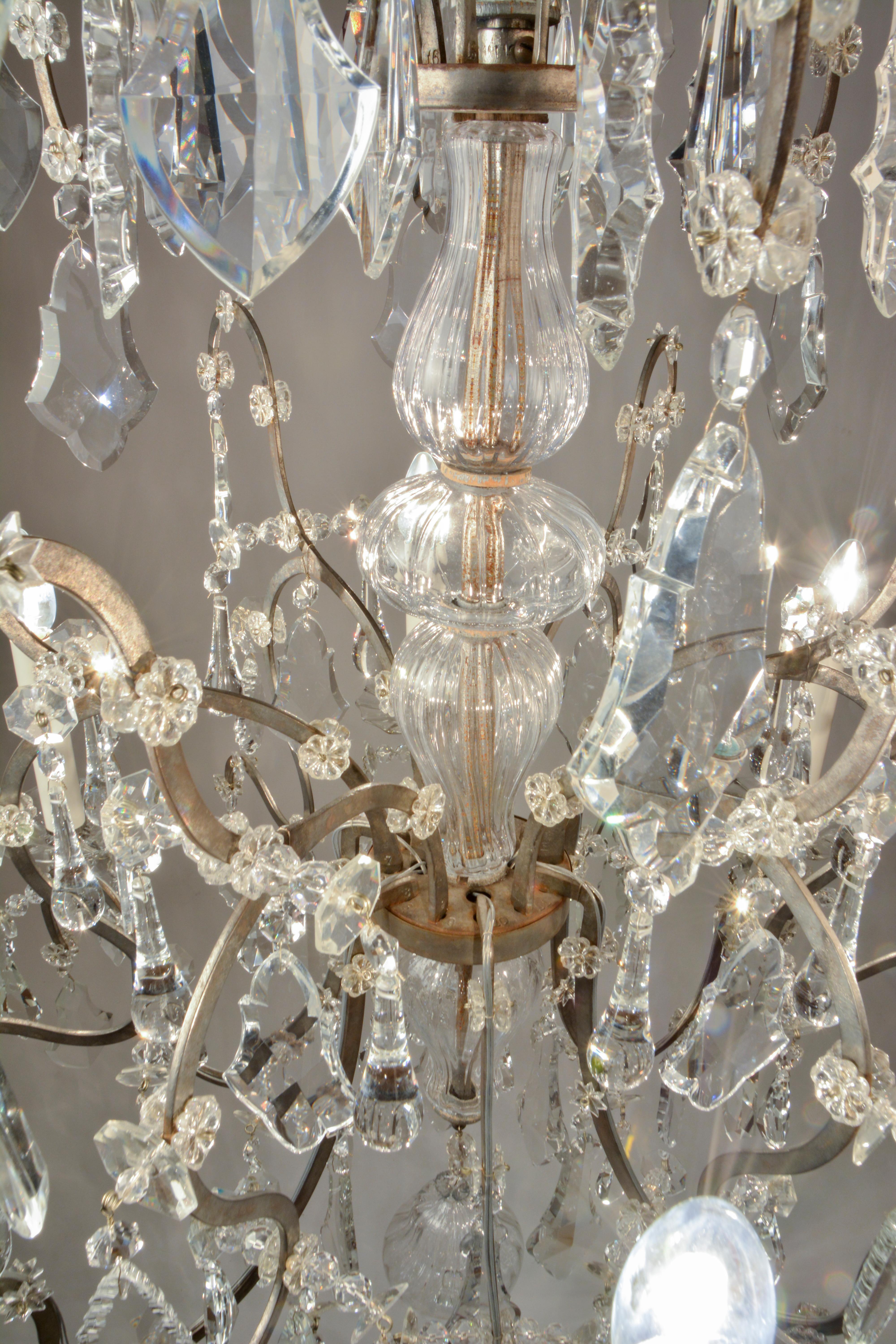 This is a wonderful example of high-Baroque wrought iron chandeliers. The frame has a protective zinc finish that is in good condition with minor fading. 
The crystals are hand-cut and polished and are complete. Missing parts have been replaced