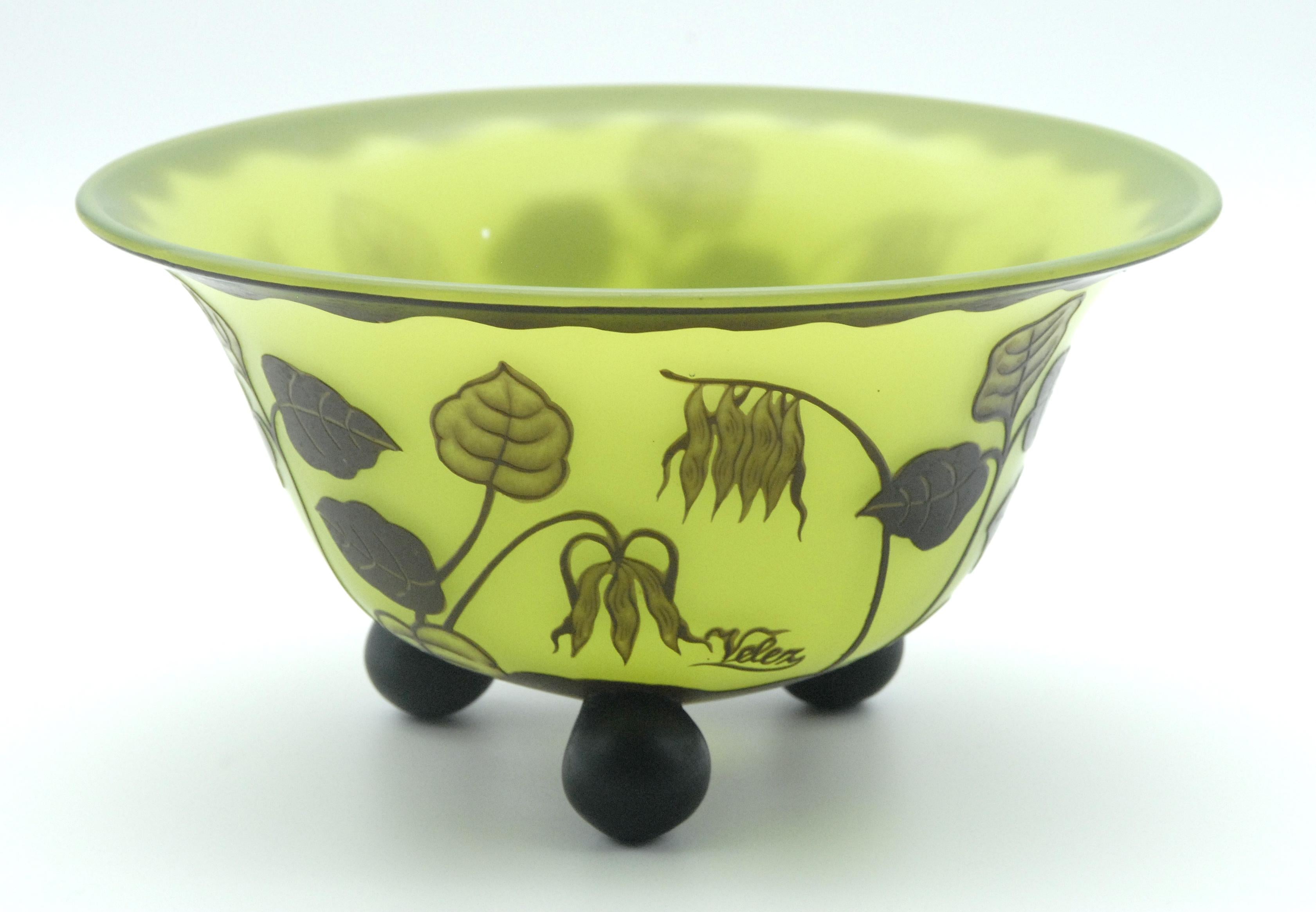 Lime green colored glass flared bowl overlaid with a topaz colored design of leafy plants and flowers resting on 3 ball feet. Signed 'Velez' in the topaz colored glass.