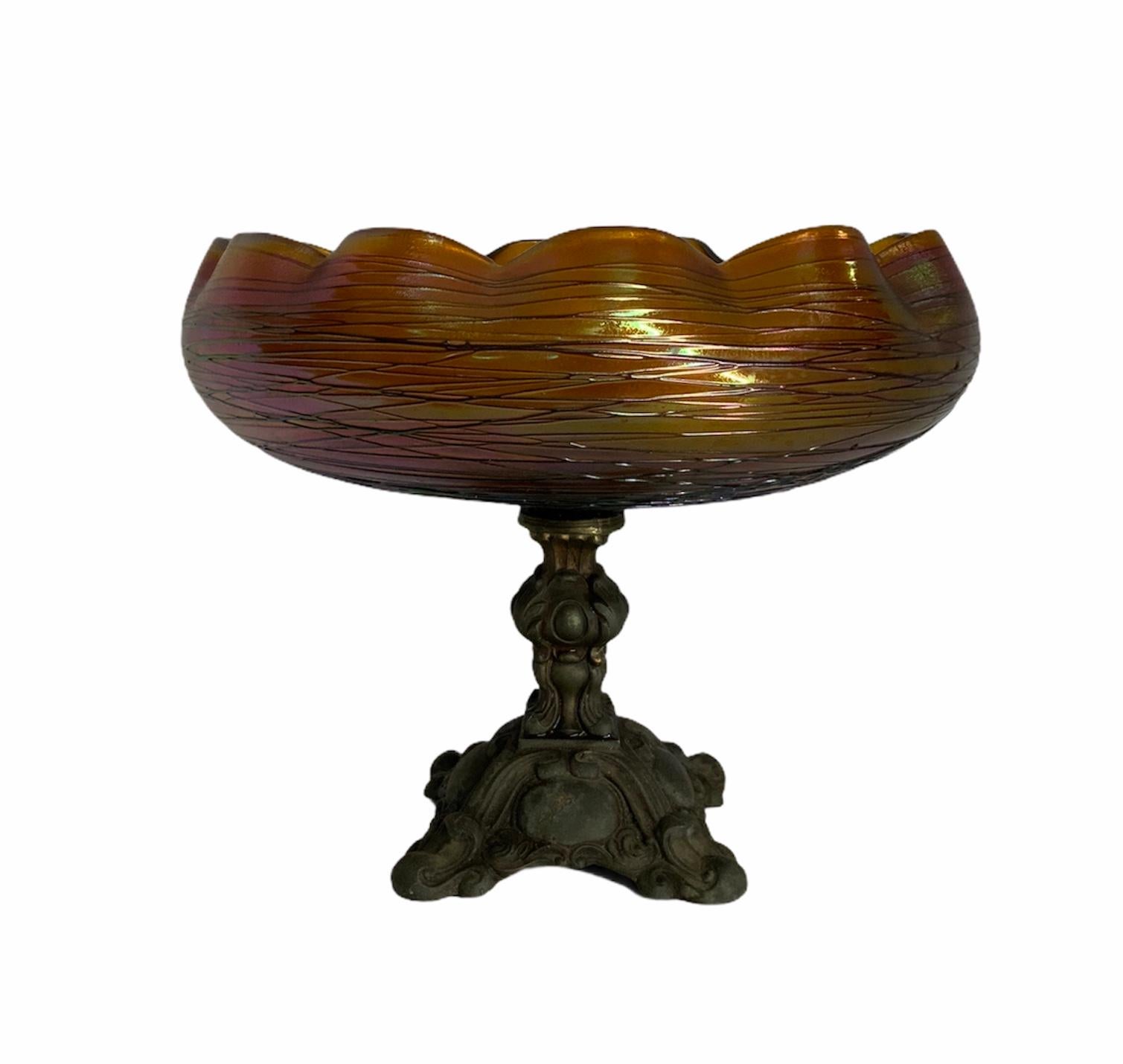 This is a threaded iridescent gold yellow-orange glass scalloped rim bronze compote/vase. The bronze pedestal is carved and shaped as a wide column ending as a quatrefoil. It is adorned with acanthus leaves and scrolls. Inside the bowl, there is an