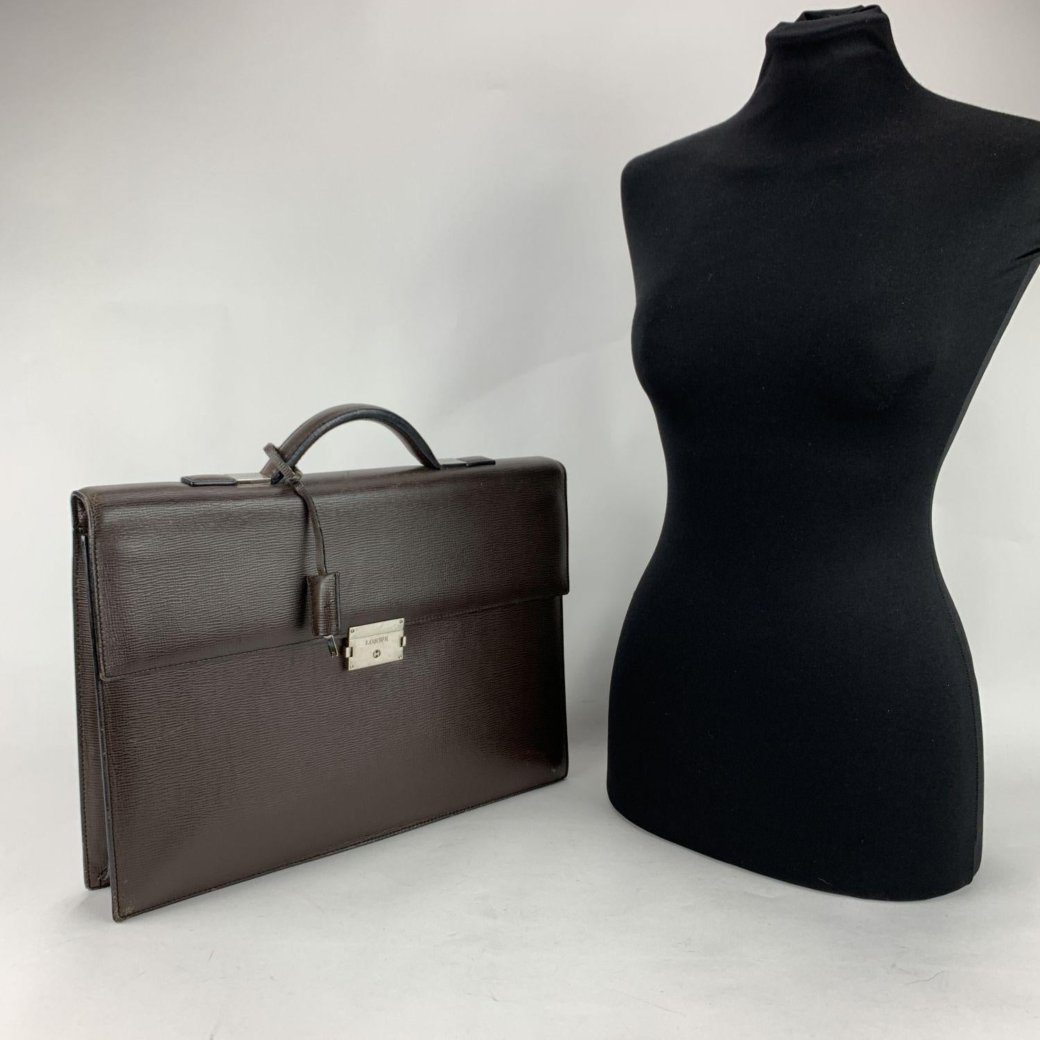 Loewe briefcase in brown textured leather. Sleek & elegant. Silver metal hardware. Single flat top handle. Flap with push lock closure with key (included). 1 flat section under the flap. 1 rear pocket. Fabric lining inside with 1 side zip pocket.