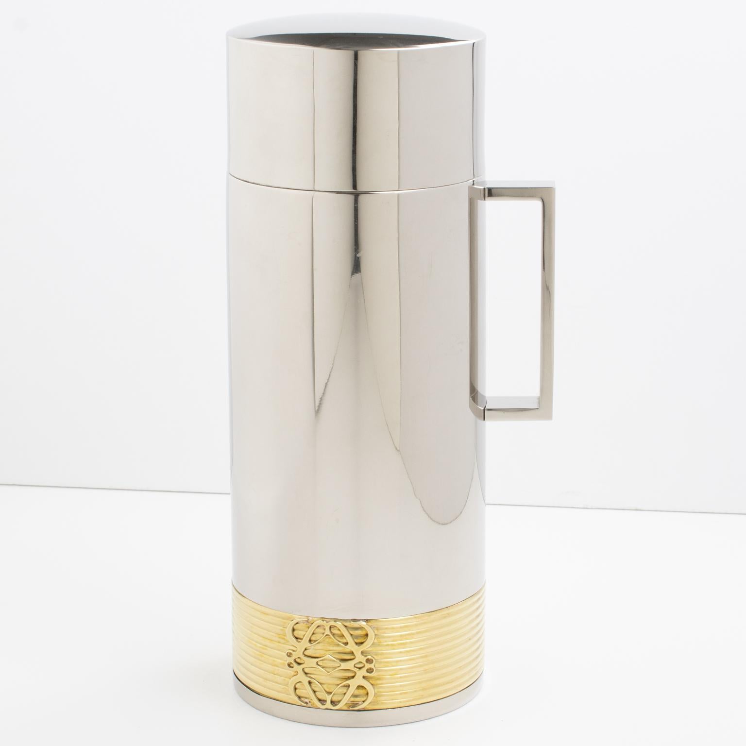 This is a sleek and modernist barware thermos made of chrome and gilded brass, produced by Loewe in Spain. The insulated decanter features a large flat handle and a dimensional striped design with a gilded brass Loewe logo decor. The inner thermos