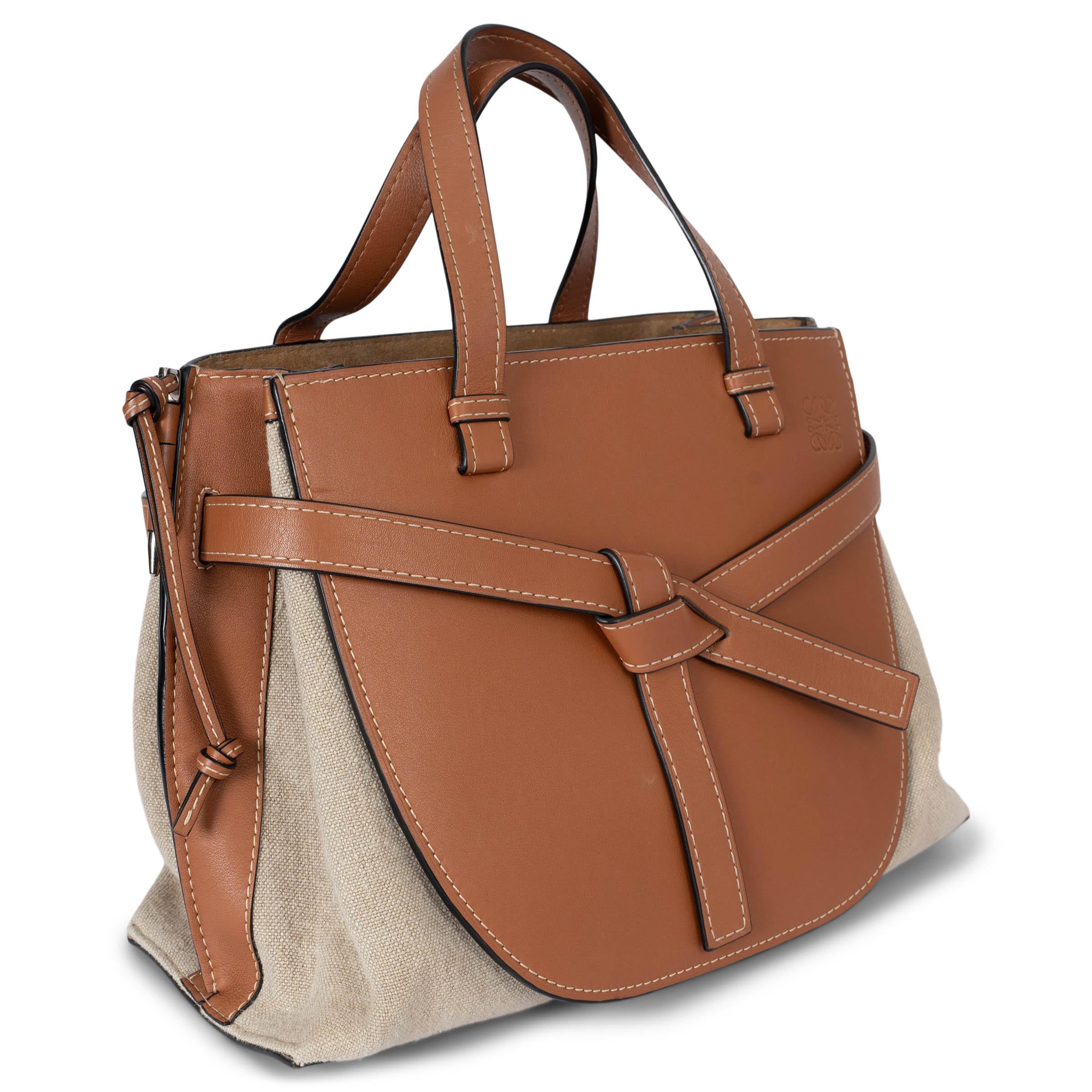 Este bolso tote Gate Medium 100% auténtico de Loewe combina piel tostada y lona avena, acentuada con pespuntes beige para un aspecto refinado y atemporal. La parte delantera luce un detalle de cinturón, e incluye una bandolera ajustable y