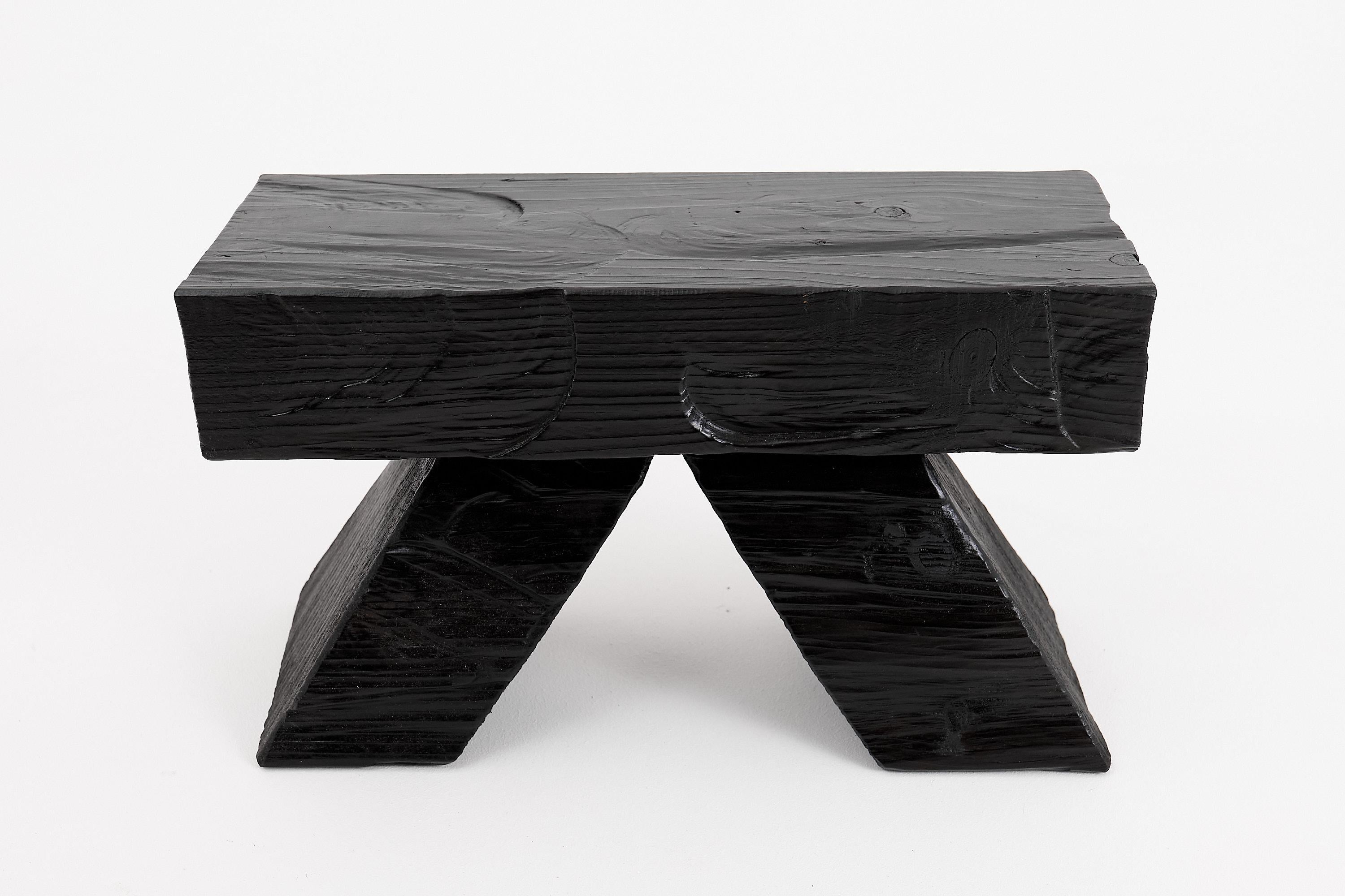 This striking blackened wood stool combines contemporary minimalism with raw, natural appeal. Expertly handcrafted, the piece features a substantial rectangular seat supported by two angular, wedge-shaped legs, creating a bold, sculptural profile.