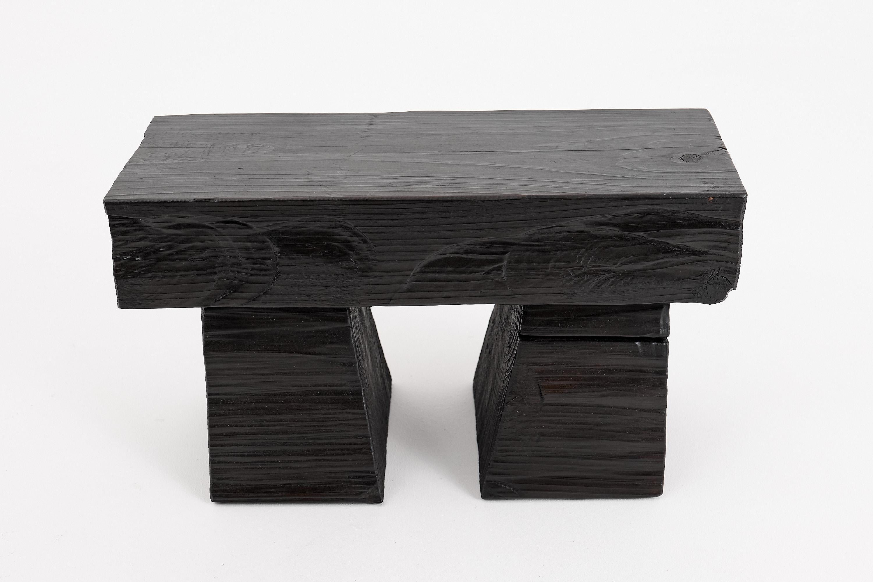 Fai colpo con questo sgabello scultoreo di grande impatto, meticolosamente realizzato in legno massiccio e rifinito con un audace effetto annerito e carbonizzato. Il pezzo presenta una sostanziale seduta rettangolare sostenuta da due robuste gambe