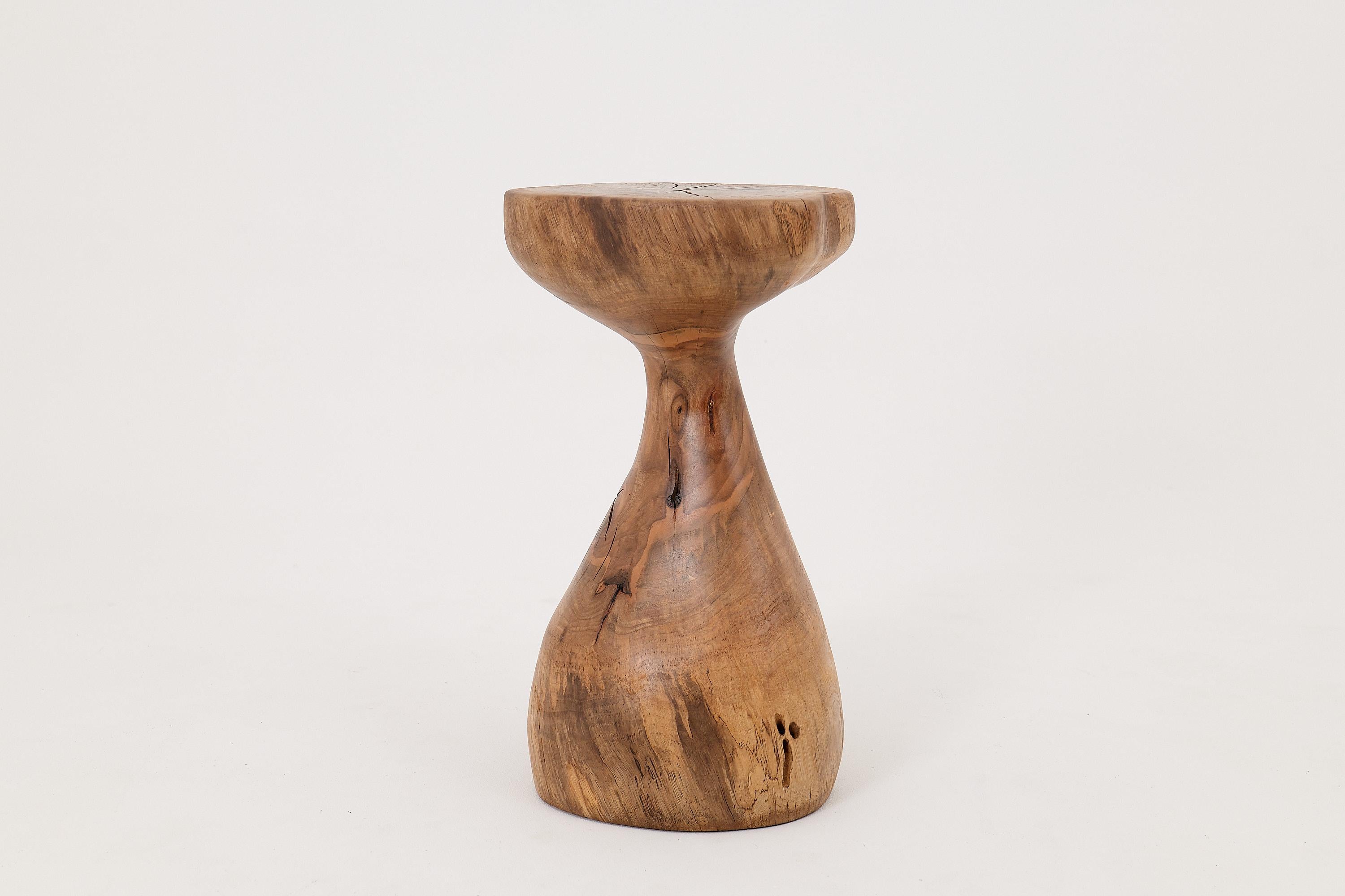 Esta escultural mesa auxiliar es una pieza única de arte funcional, tallada totalmente a mano a partir de un único tronco de madera dura. Su forma se eleva desde una base ancha y asentada que se estrecha con gracia antes de ensancharse de nuevo en