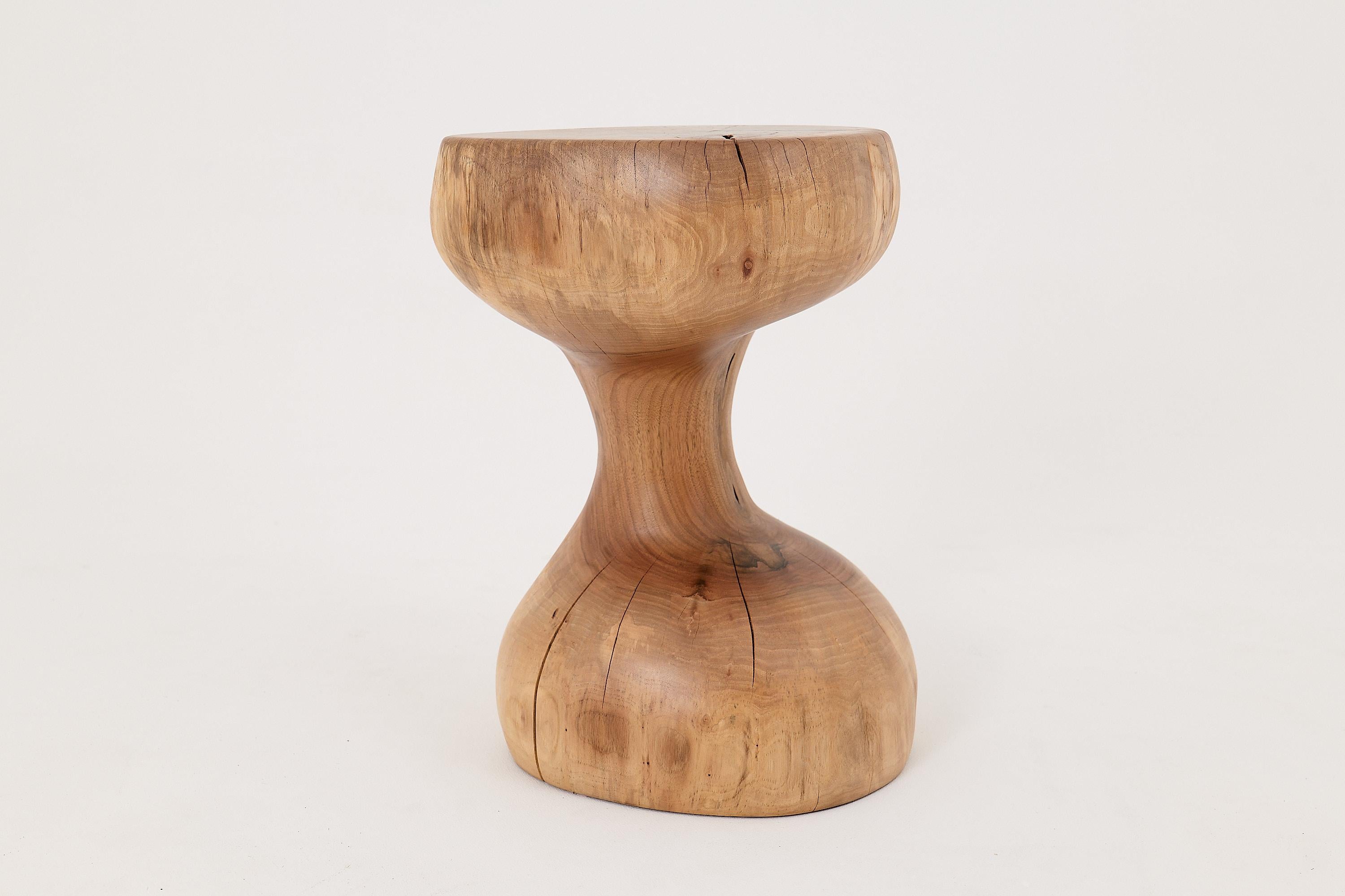Esta escultural mesa auxiliar es una obra de arte funcional única en su género, tallada totalmente a mano a partir de un único tronco de madera dura. Su silueta, similar a la de un reloj de arena, fluye a la perfección desde una base redondeada