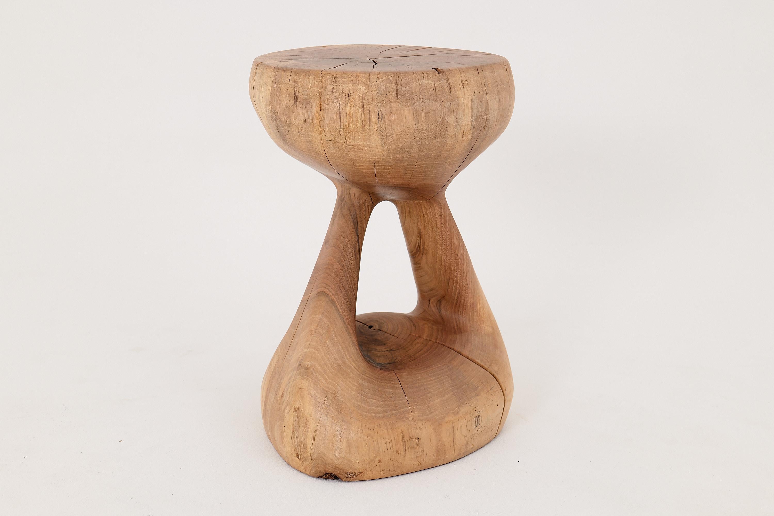 Esta escultural mesa auxiliar es una pieza única, tallada a mano a partir de un único tronco de madera maciza. Su forma recuerda a un reloj de arena en movimiento, con la parte superior y la base acampanadas conectadas por un centro esbelto y