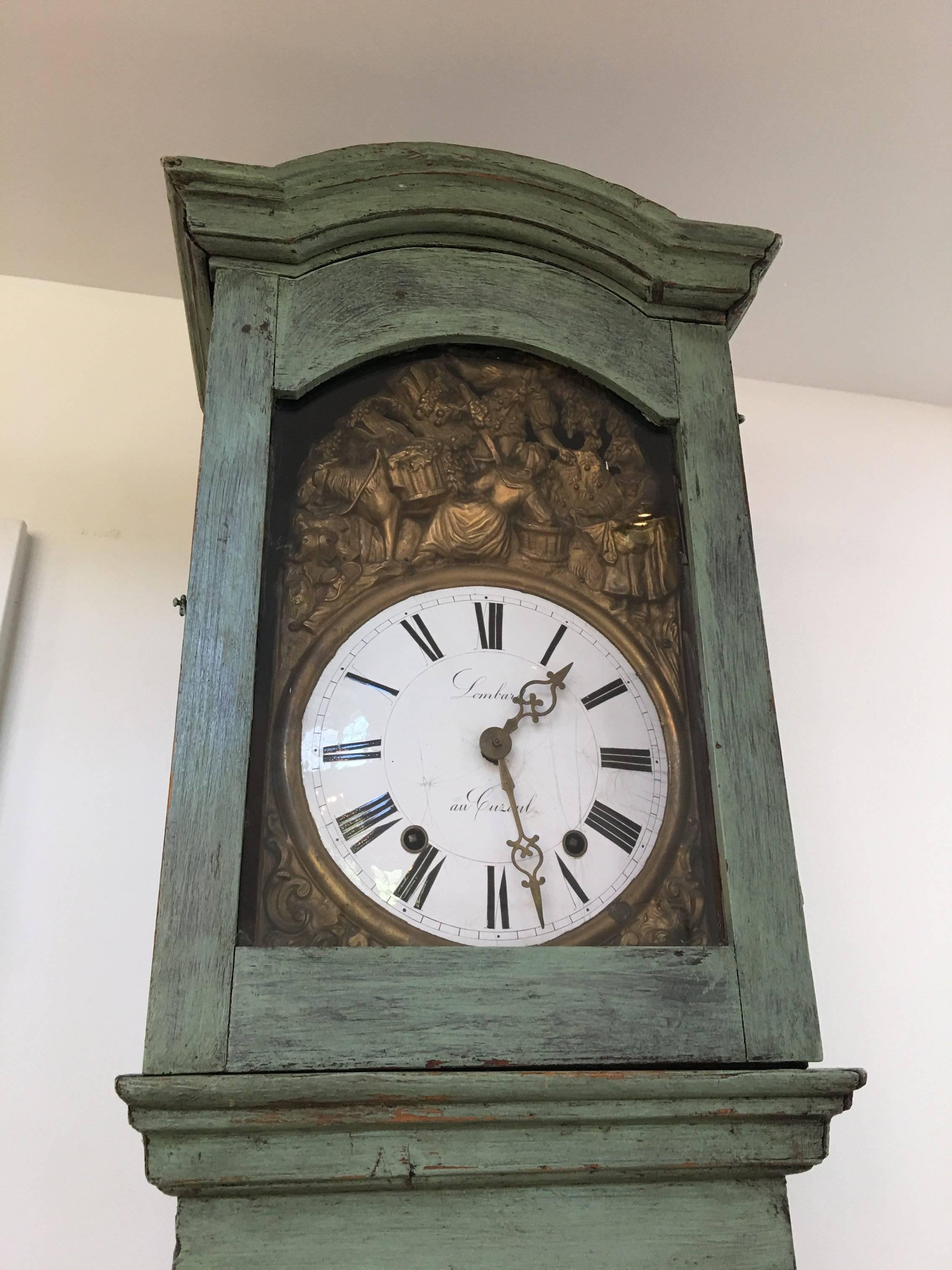 Antique Lombard au Cuzoul grandfather clock. Green-washed wood with intricate brass detailing around white face and wooden door on hinges.