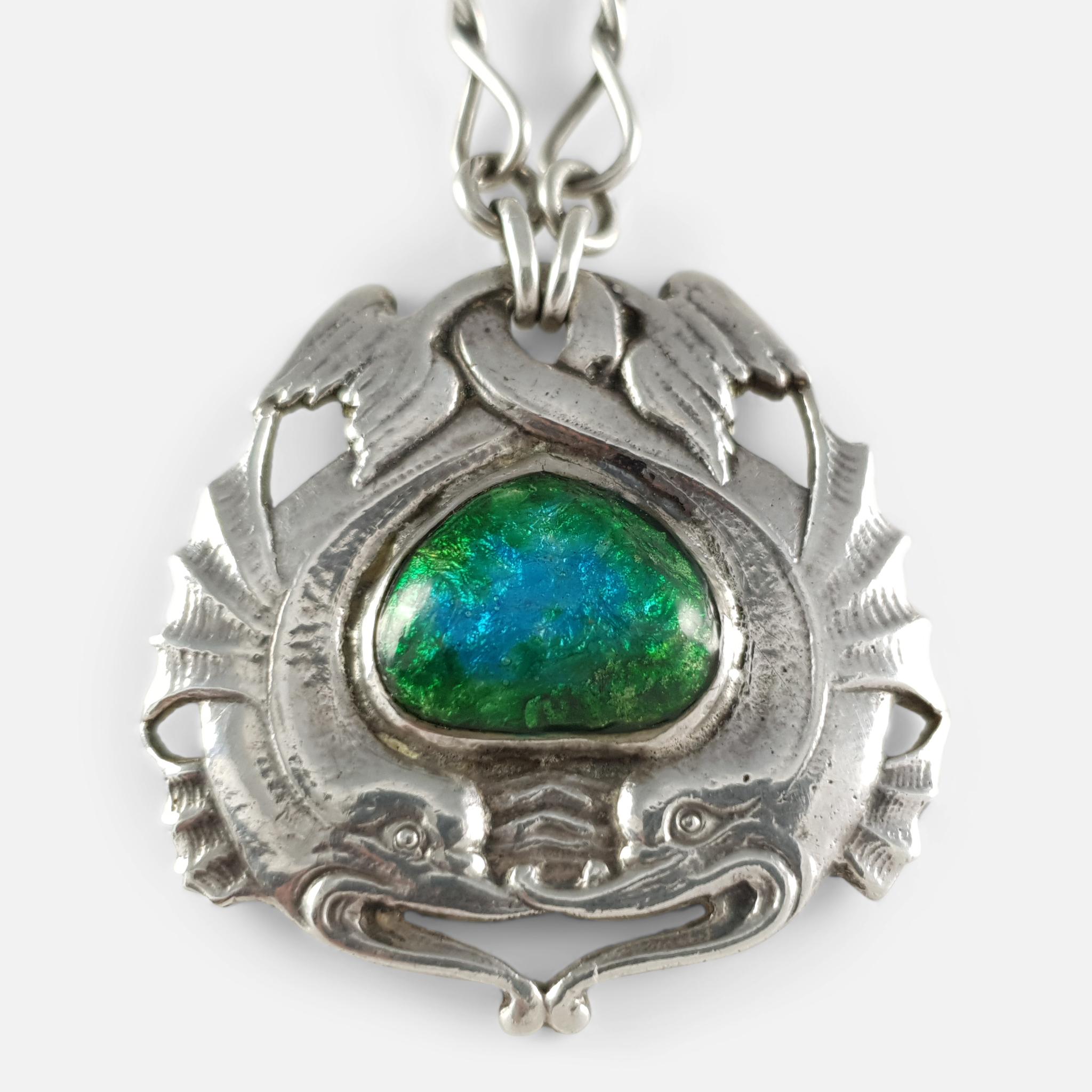 An Arts and Crafts ornamental sterling silver necklace by Ramsden & Carr. The necklace is designed as two intertwined mythical sea creatures encircling a central blue and green enamel plaque, on silver twisted-link chain, and a shield-shaped silver
