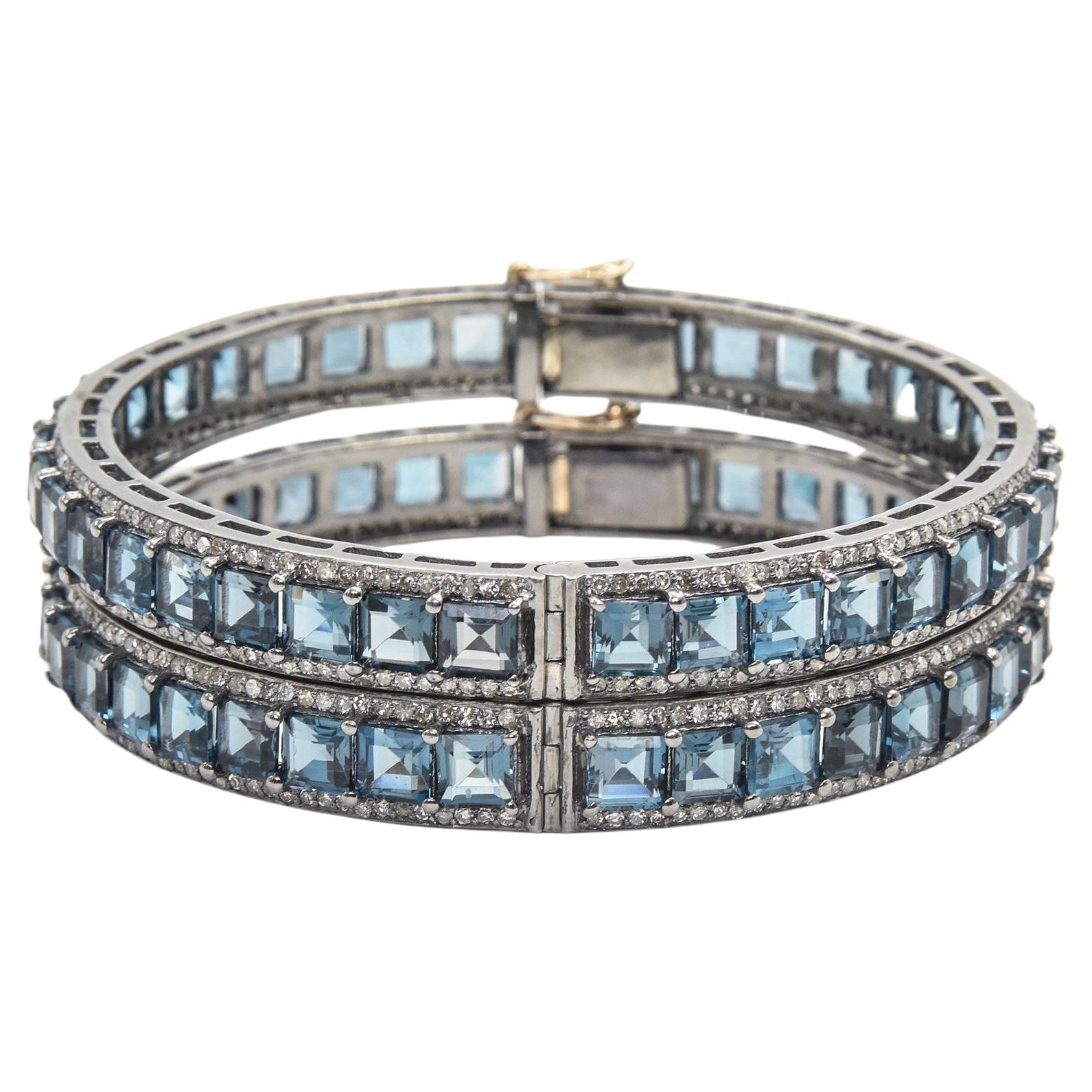 Pair of London blue topaz and diamond bangles mounted in sterling silver with a 14k gold tongue clasp and safety. Each bracelet contains princess cut blue topaz prong set down the center with round diamonds along the side edges.  The 2 bracelets