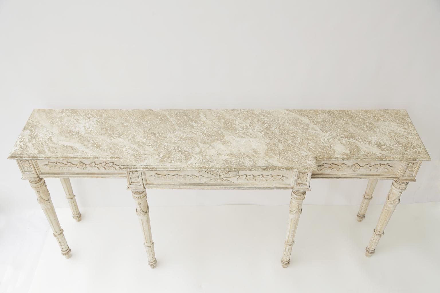 Console table, long and narrow proportions, having a faux painted breakfront top, in Louis XVI taste, on table with painted finish showing natural wear, its fielded apron out carved with foliate carving, raised on round stop-fluted leg, decorated