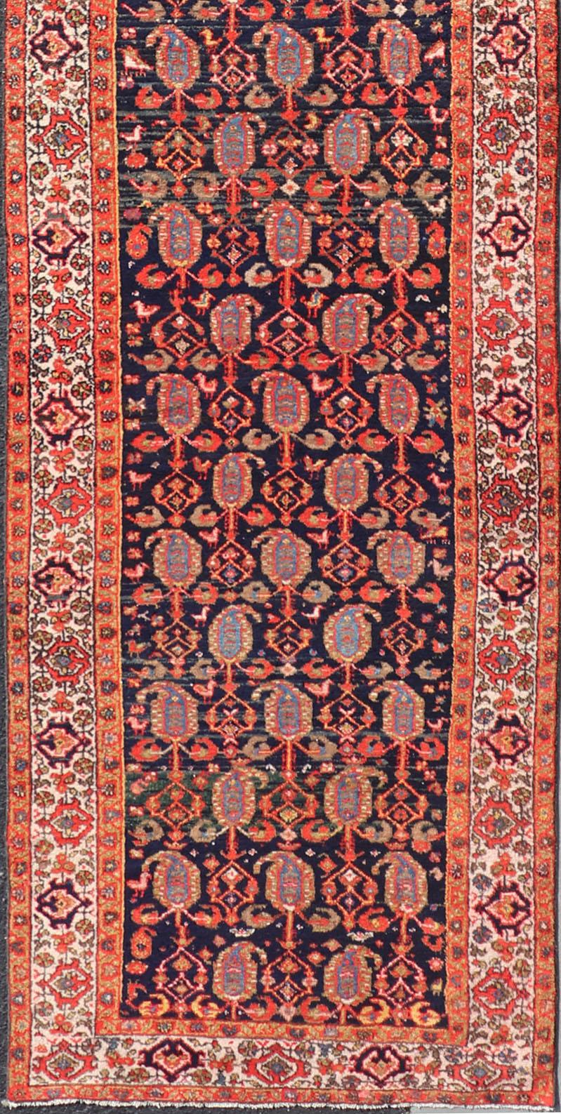 Measures: 3'3 x 15'2.

This 1920's Persian Hamadan runner features vivid shades of navy, blue, red-orange, green and small accents and ivory. The field design is a unique paisley motif design scattering across the entirety of the field. The border