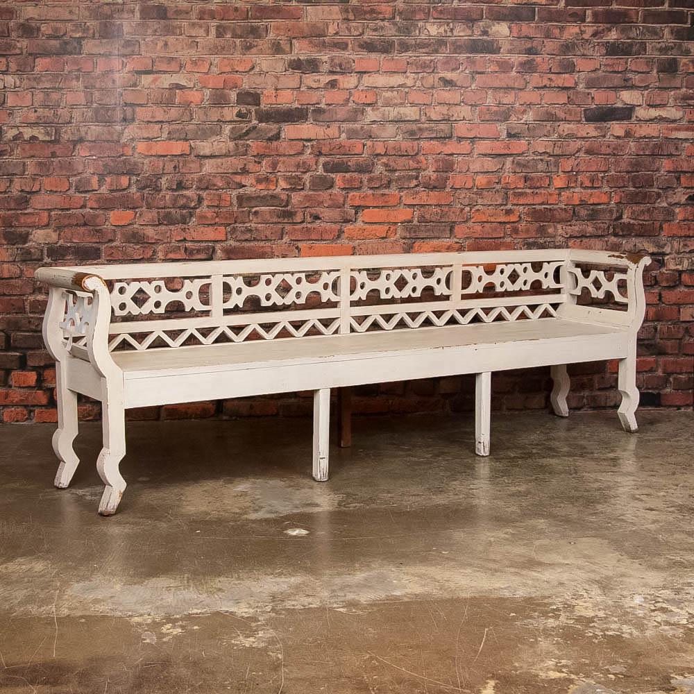 There is something whimsical about this long Swedish bench with it's pierced back splat and repetitive triangle design reminiscent of a fairy tale. The distressed off white paint with wear on the arms exemplifies a rustic, country charm. The pegged
