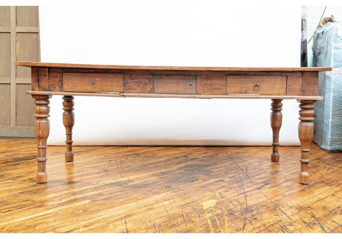 Ca. 18th-19th century A long wood farm table with an overhanging three plank top constructed with dowels over a frieze with two drawers one on each end and one hinged drawer in the center. Two drawers are lacking the brass ring pulls. The entire