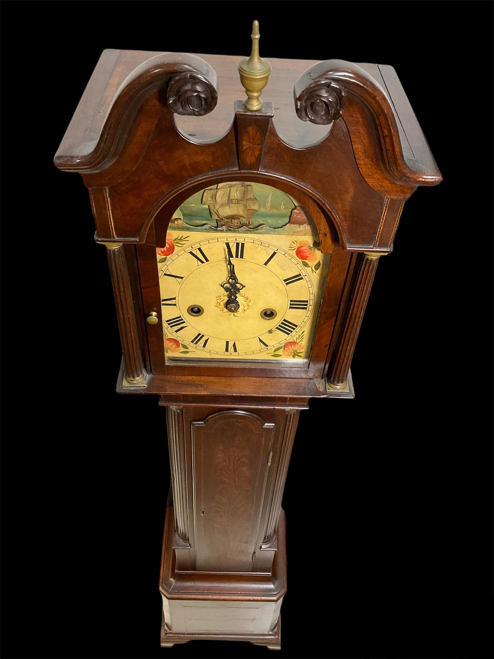 This is a long case Dwarf wood clock. Its bonnet is decorated with a swan neck pediment at the top and an a bronze urn shaped finial. Below the finial, there is an inlaid paterae. The front of the bonnet is also embellished by reeded columns with